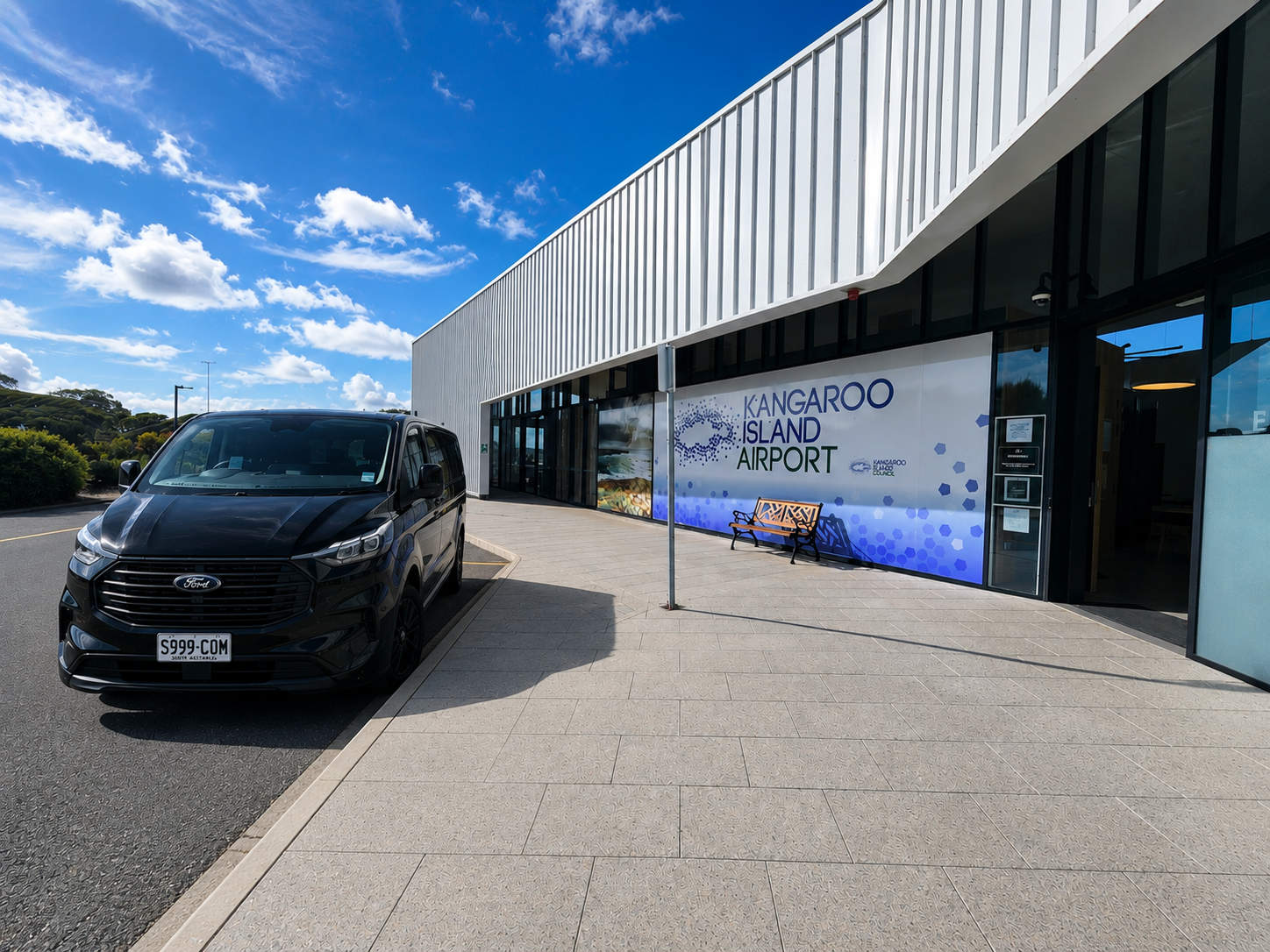 Exterior of Kangaroo Island Airport with black Ford vehicle parked outside, clear blue sky with scattered clouds, and a bench near the entrance.