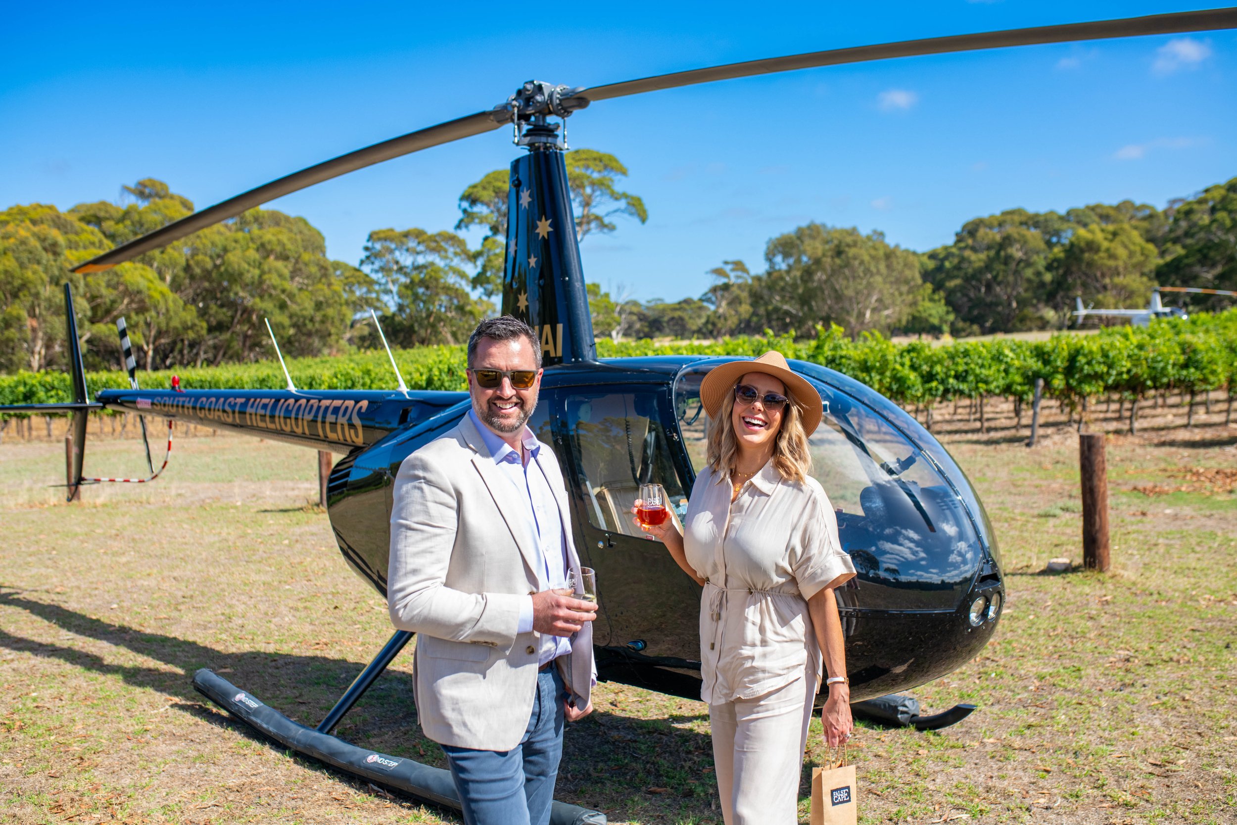 Two people, a man and a woman, standing in front of a black helicopter in a vineyard, holding glasses of wine, smiling, with a helicopter and green trees in the background on a sunny day.