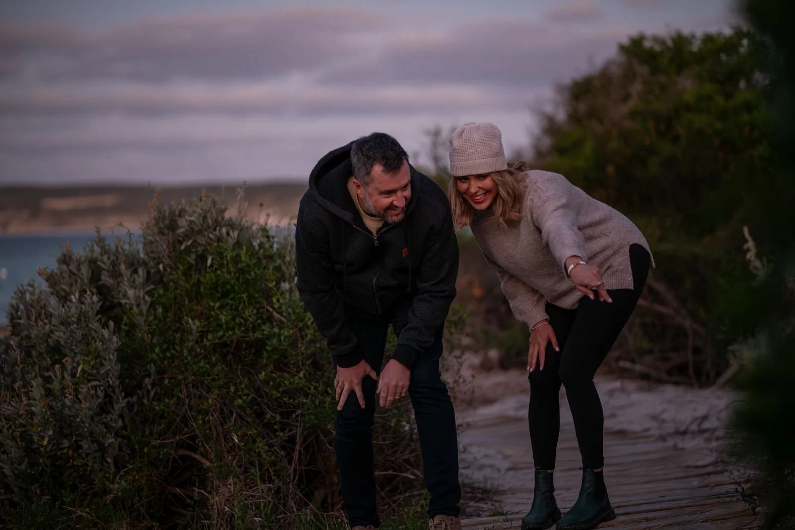 A man and woman smiling and crouching down near bushes and a wooden pathway outdoors during dusk, looking at Little Penguins under bushes.
