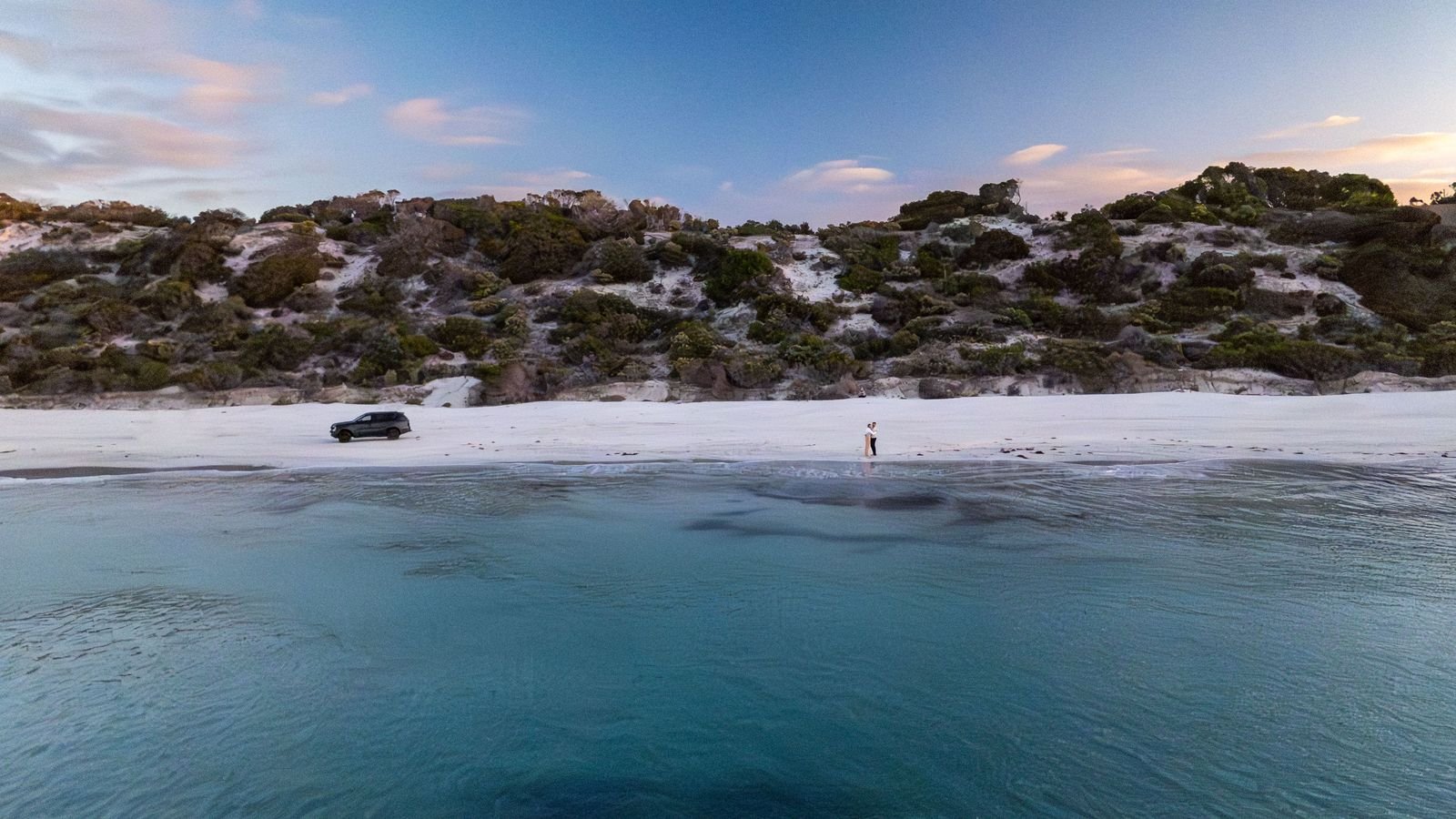 Emu Bay beach with a white sand shoreline, a car parked on the sand, and a person standing near the water's edge. Behind the beach is a rocky, shrub-covered hillside under a partly cloudy sky.