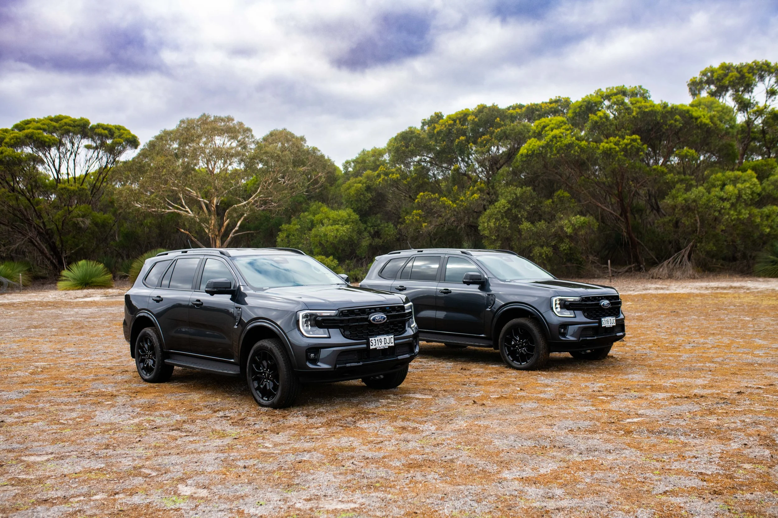 Two black SUVs parked on a dirt area with grass and trees in the background under a cloudy sky.