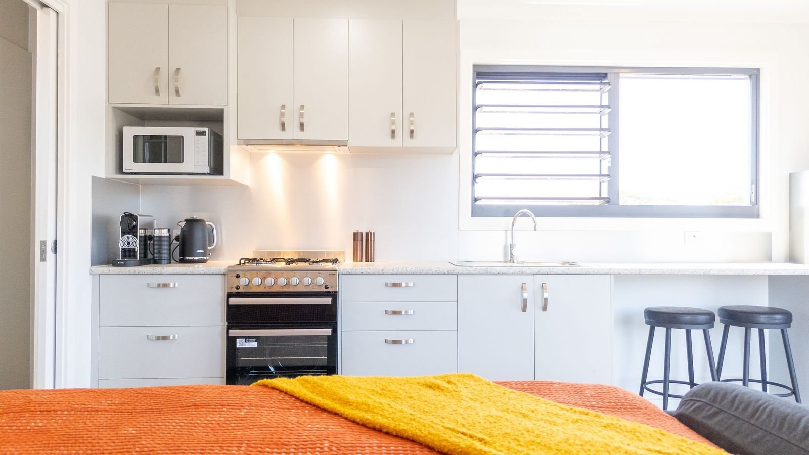 Bright kitchen with white cabinets, a stove, microwave, coffee maker, kettle, and window with jalousie vents.