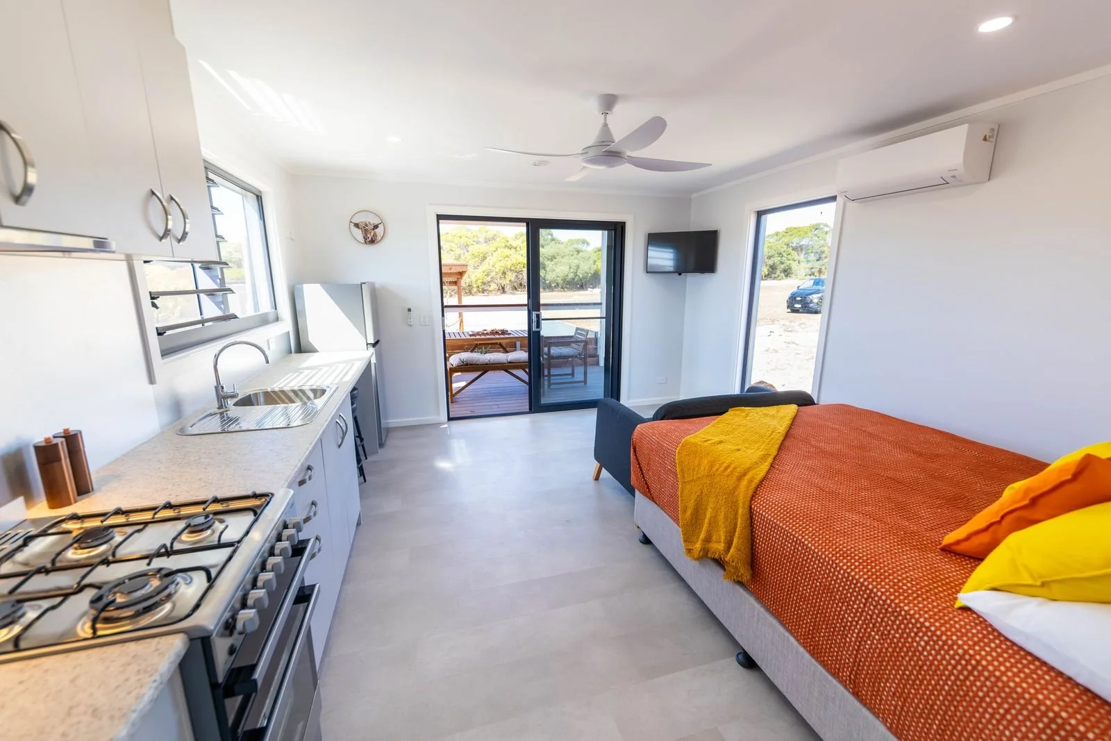 A compact living space with a small kitchen, a sofa bed with orange bedding, windows, a sliding glass door leading to a porch, and a wall-mounted TV.