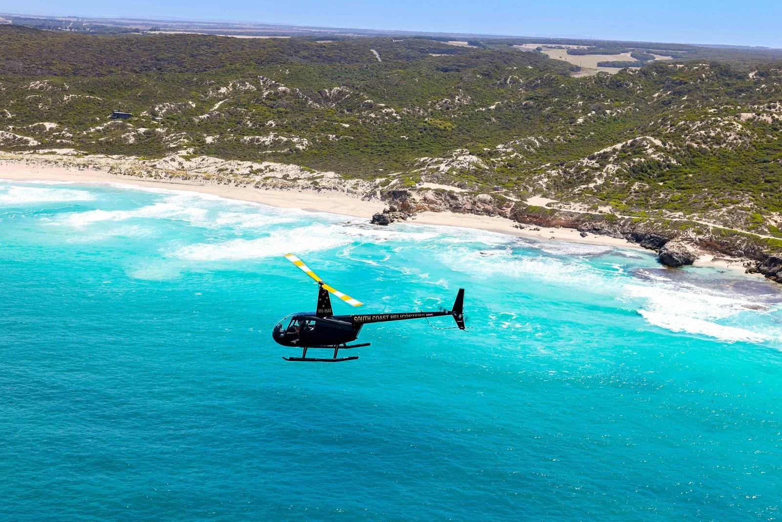 A helicopter flying over a blue ocean near a sandy beach with green, hilly terrain in the background on Kangaroo Island.