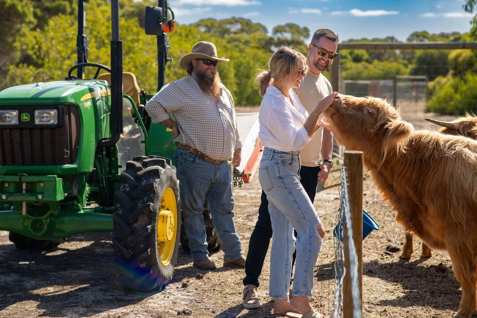 A group of three people on a farm, one woman and two men, petting a large brown cow near a wooden fence. A green tractor is parked nearby on a sunny day with a blue sky and trees in the background.