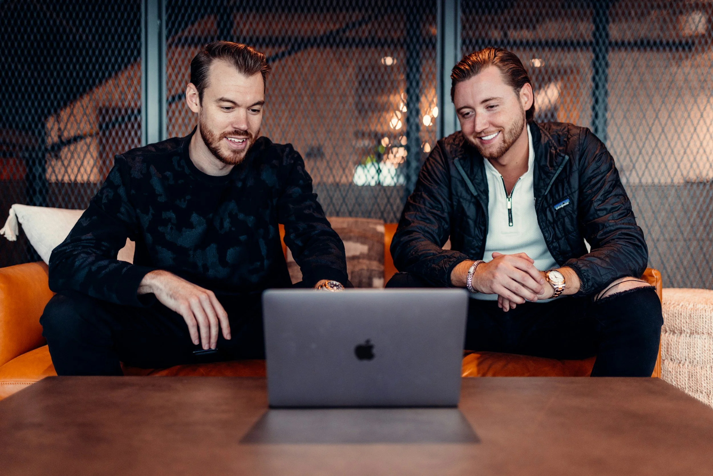 Two men sitting on a couch looking at a laptop, smiling and smiling, in a modern interior space.