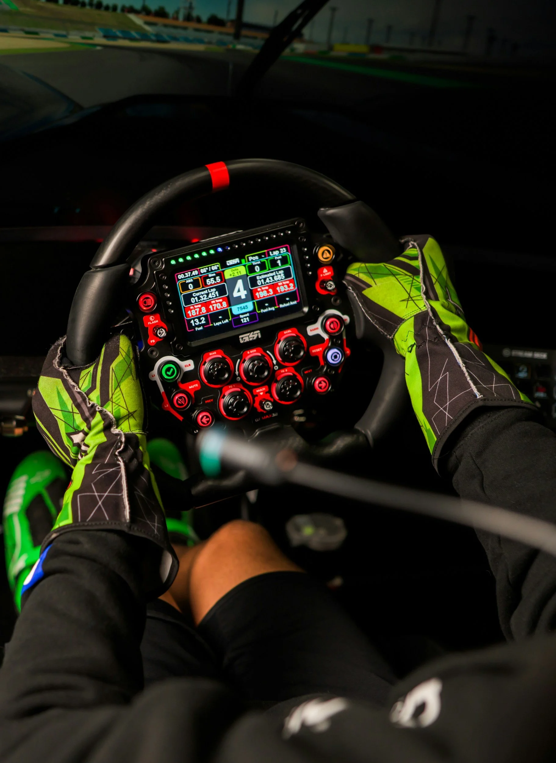 View of a race car driver's hands on a steering wheel with a digital dashboard display, wearing racing gloves and black shorts, racing on a track at night.