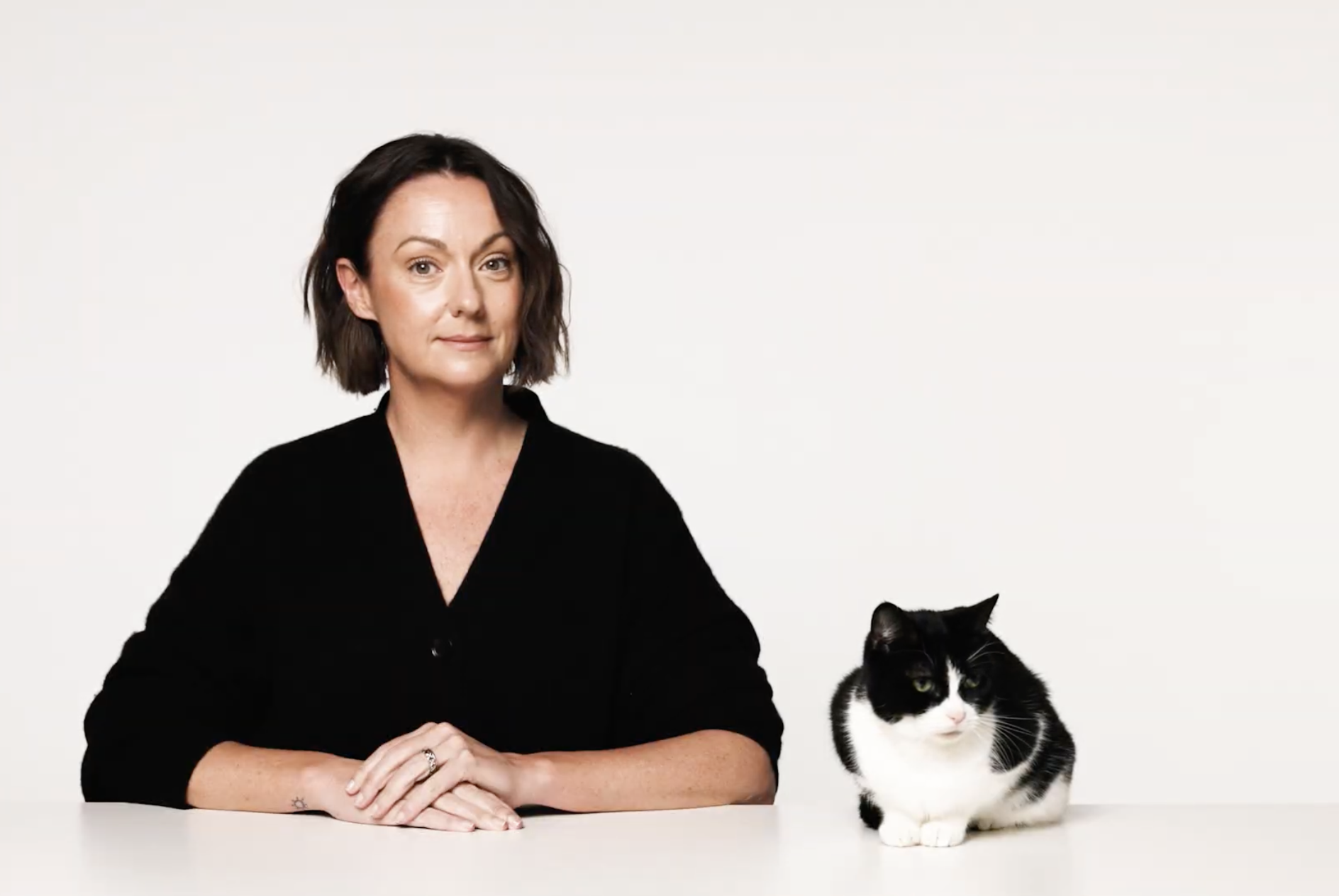 A woman with dark hair and a black top sitting at a white table with a black and white cat beside her, against a plain white background.