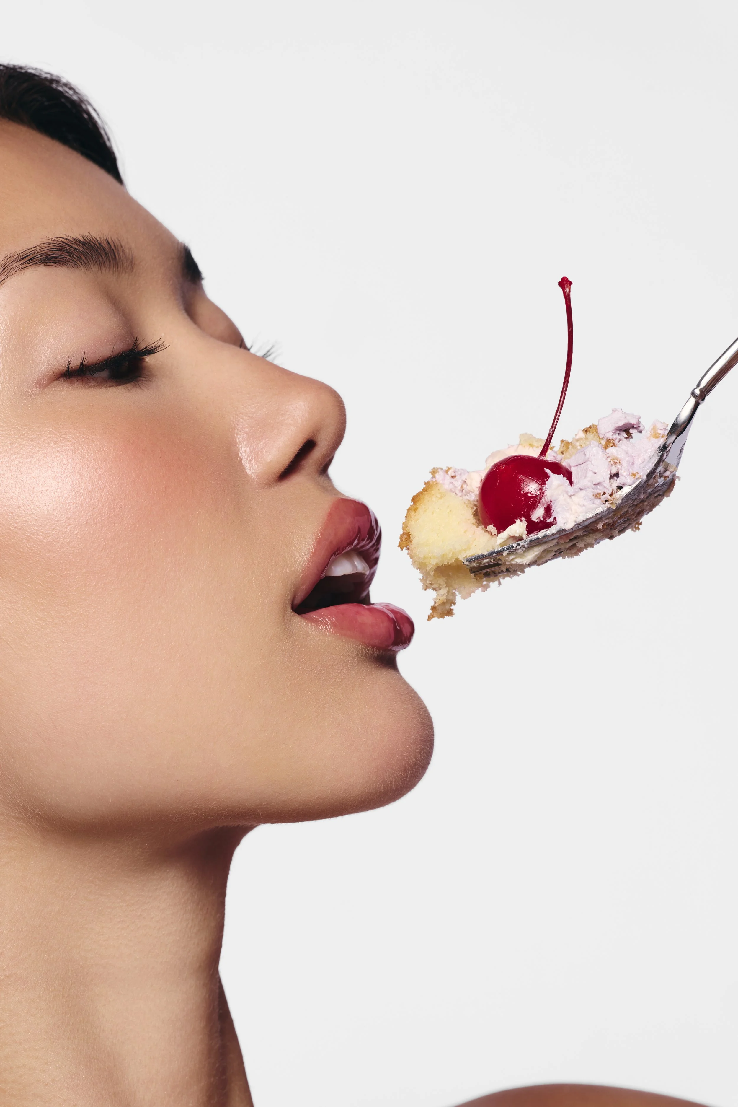 Close-up of a woman with sealed lips and makeup, about to eat a slice of cake with pink frosting, cherry, and whipped cream on a spoon.