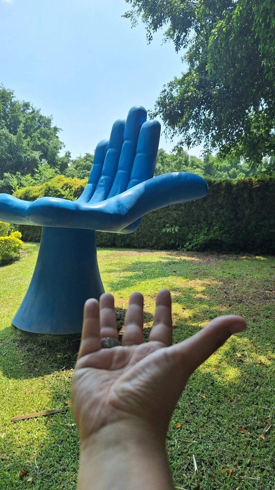 Mano extendida frente a una escultura de una mano gigante de color azul en un jardín con árboles y césped.