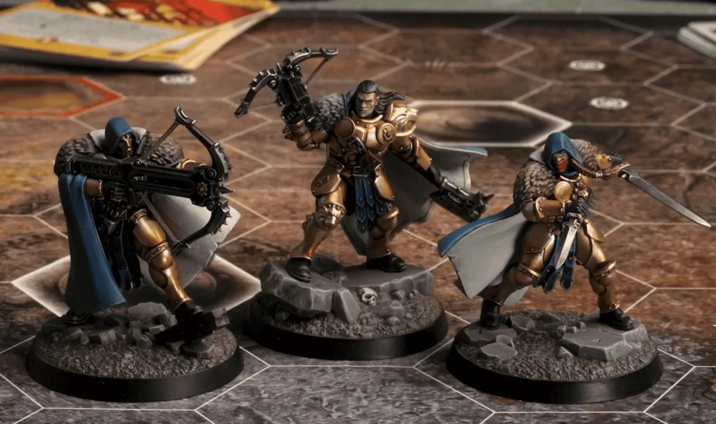 Three painted miniature figures of fantasy warriors on a game board. Each figure is on a round base with rocky terrain. The warriors are equipped with weapons and armor, and are positioned in dynamic stances.