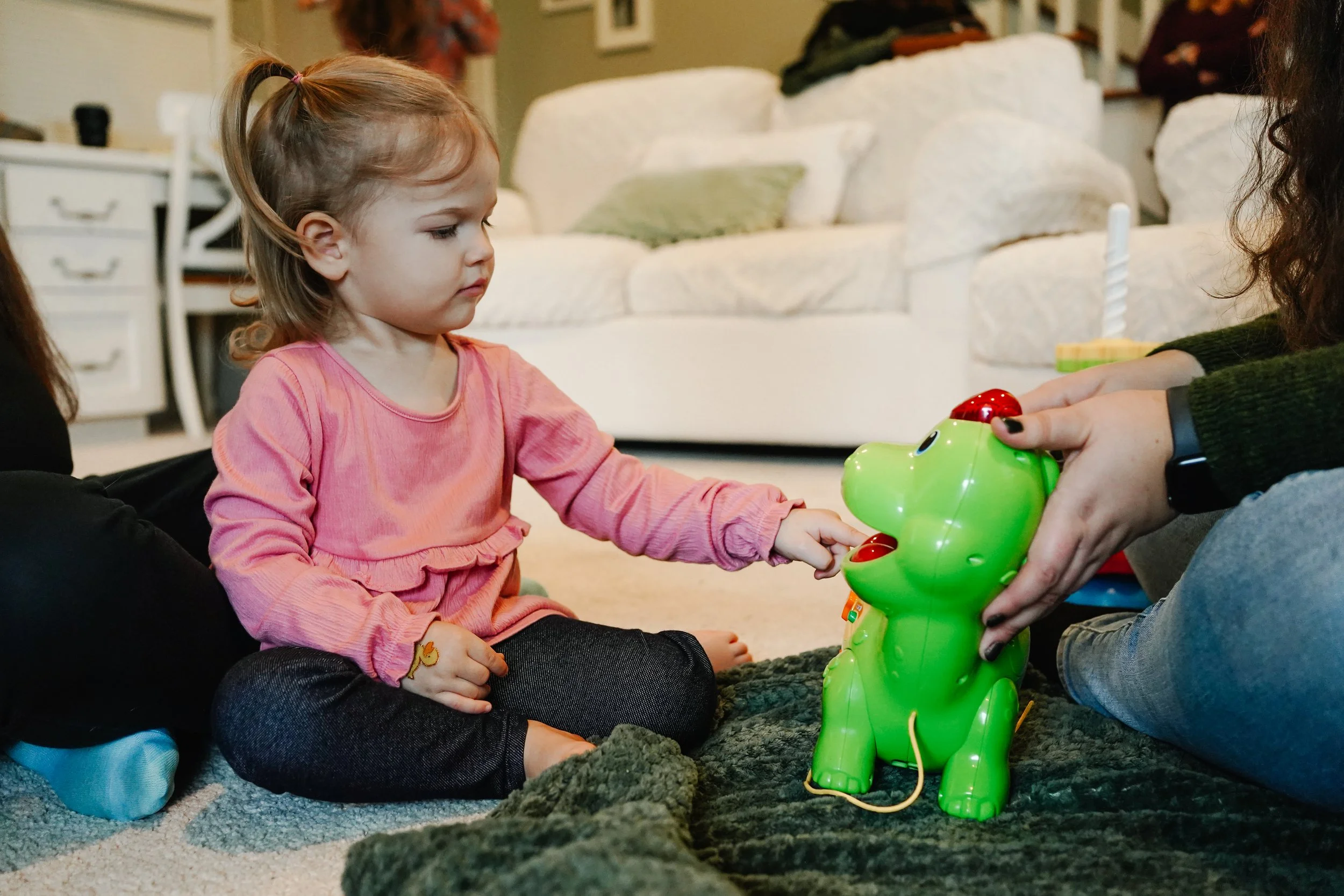 A young girl in a pink shirt and dark pants playing a green electronic toy dinosaur with a woman on a beige carpet in a living room.