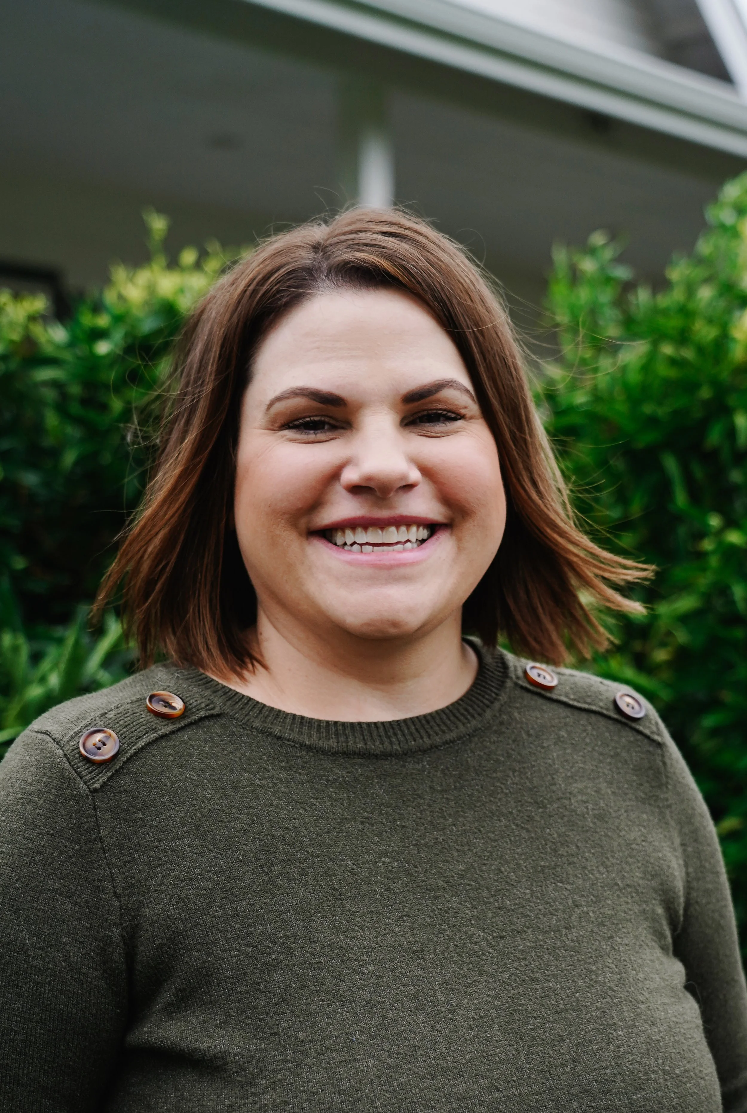 A woman with shoulder-length brown hair smiling outdoors in front of green bushes, wearing a dark green sweater with buttons on the shoulders.