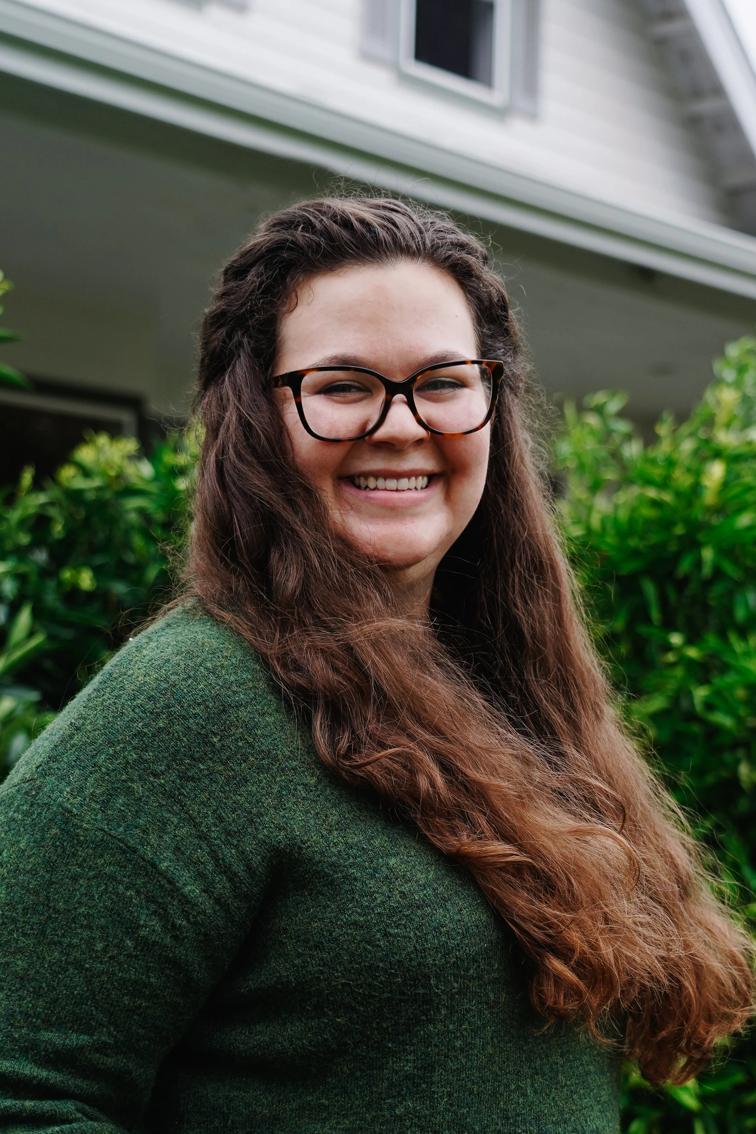 A smiling woman with long, wavy brown hair wearing glasses and a green sweater, standing outdoors in front of green bushes and a house.