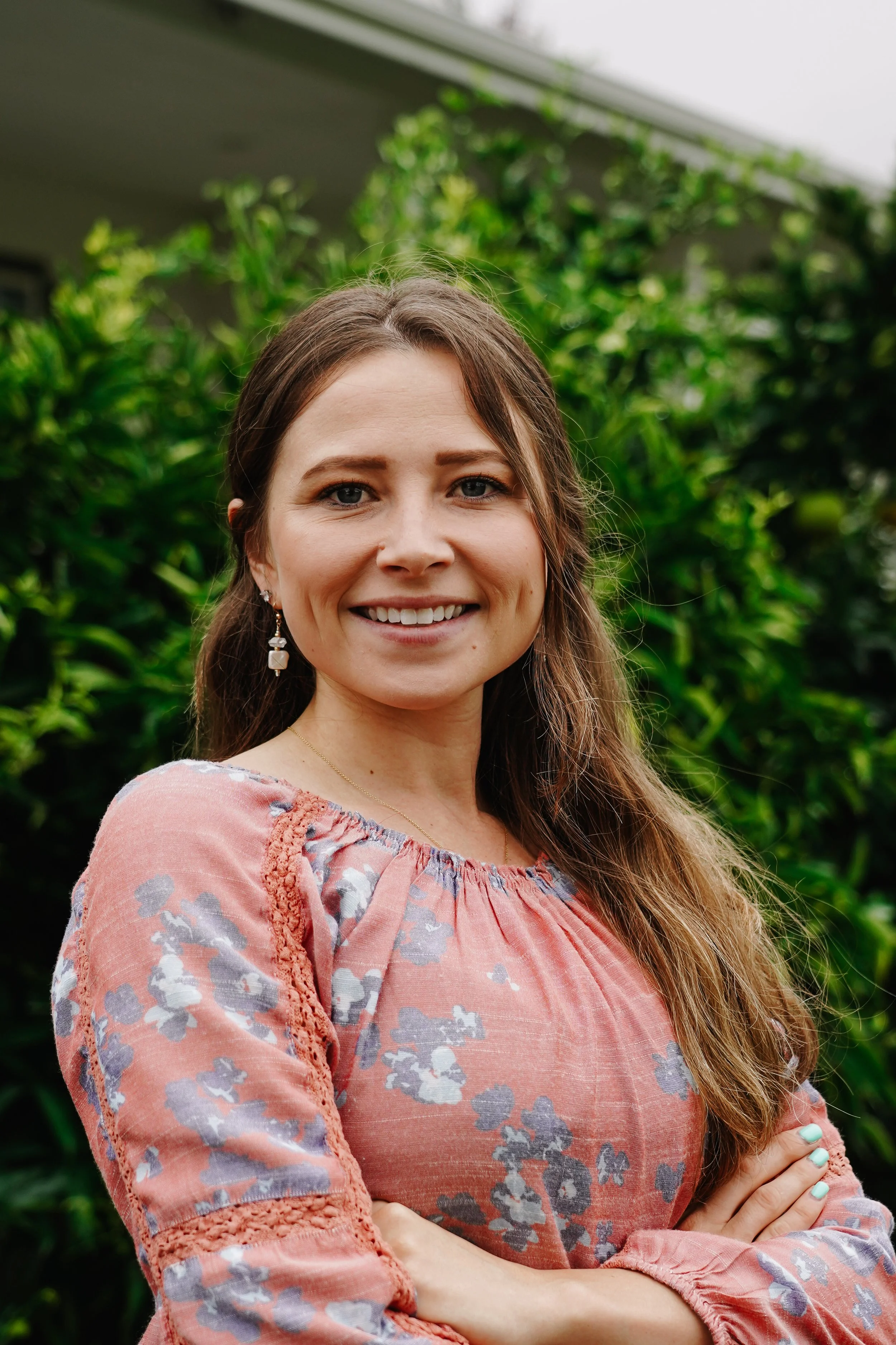 A smiling woman with long brown hair wearing a pink floral blouse standing in front of green foliage.