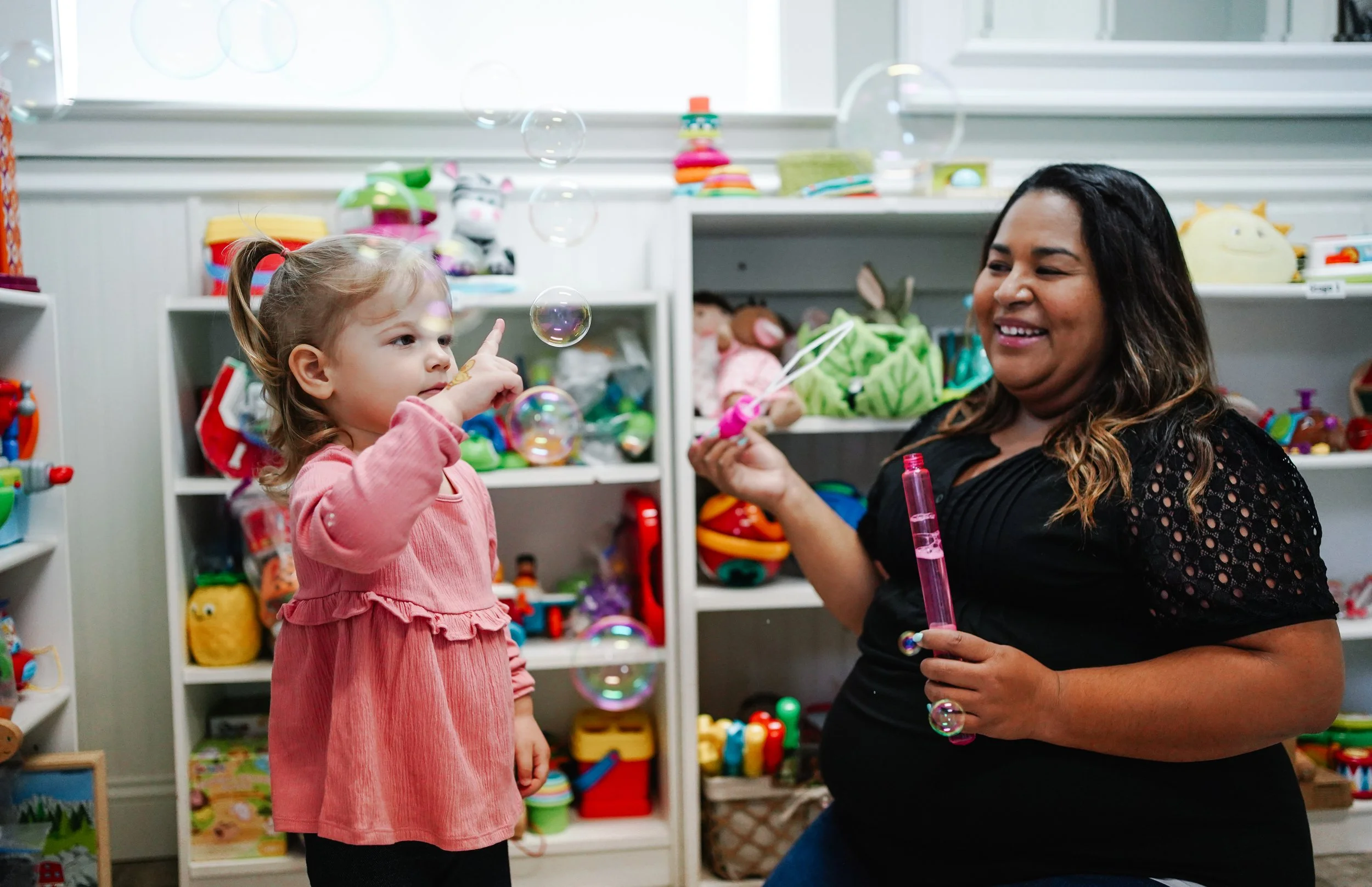 A woman and a young girl playing with bubbles in a toy room filled with colorful toys and plush animals.