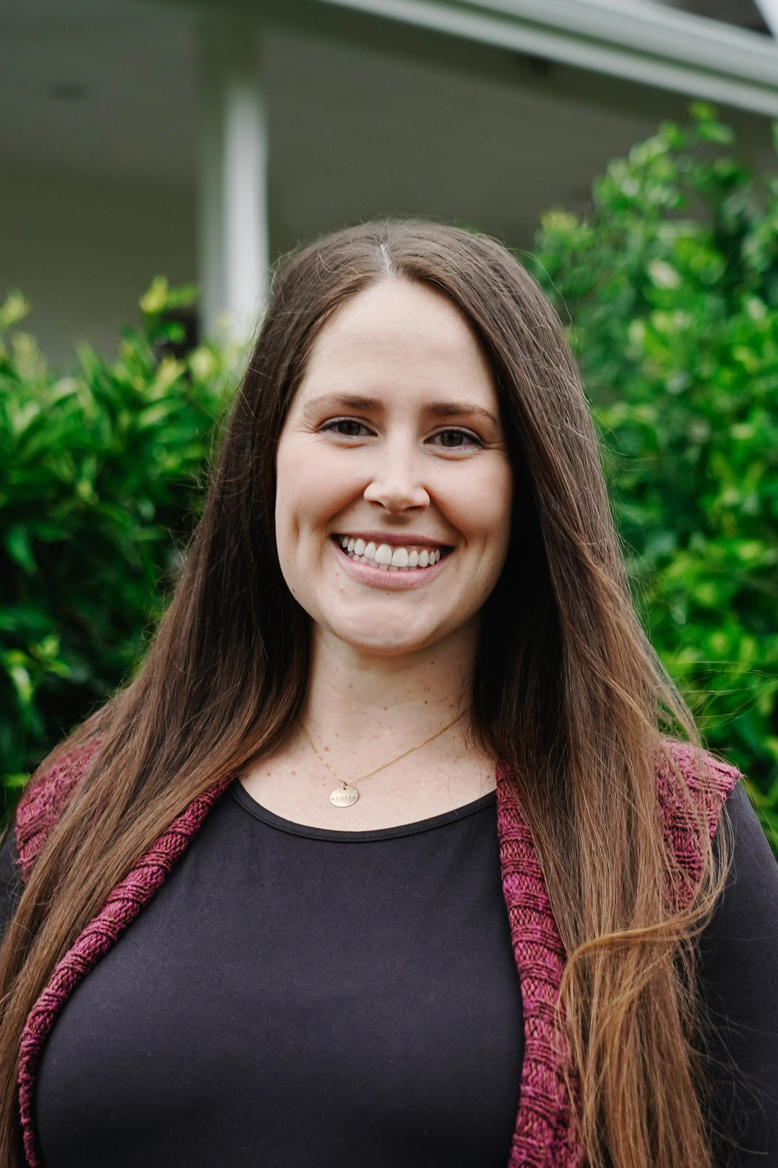 A young woman smiling outdoors, with green foliage and part of a house visible in the background.