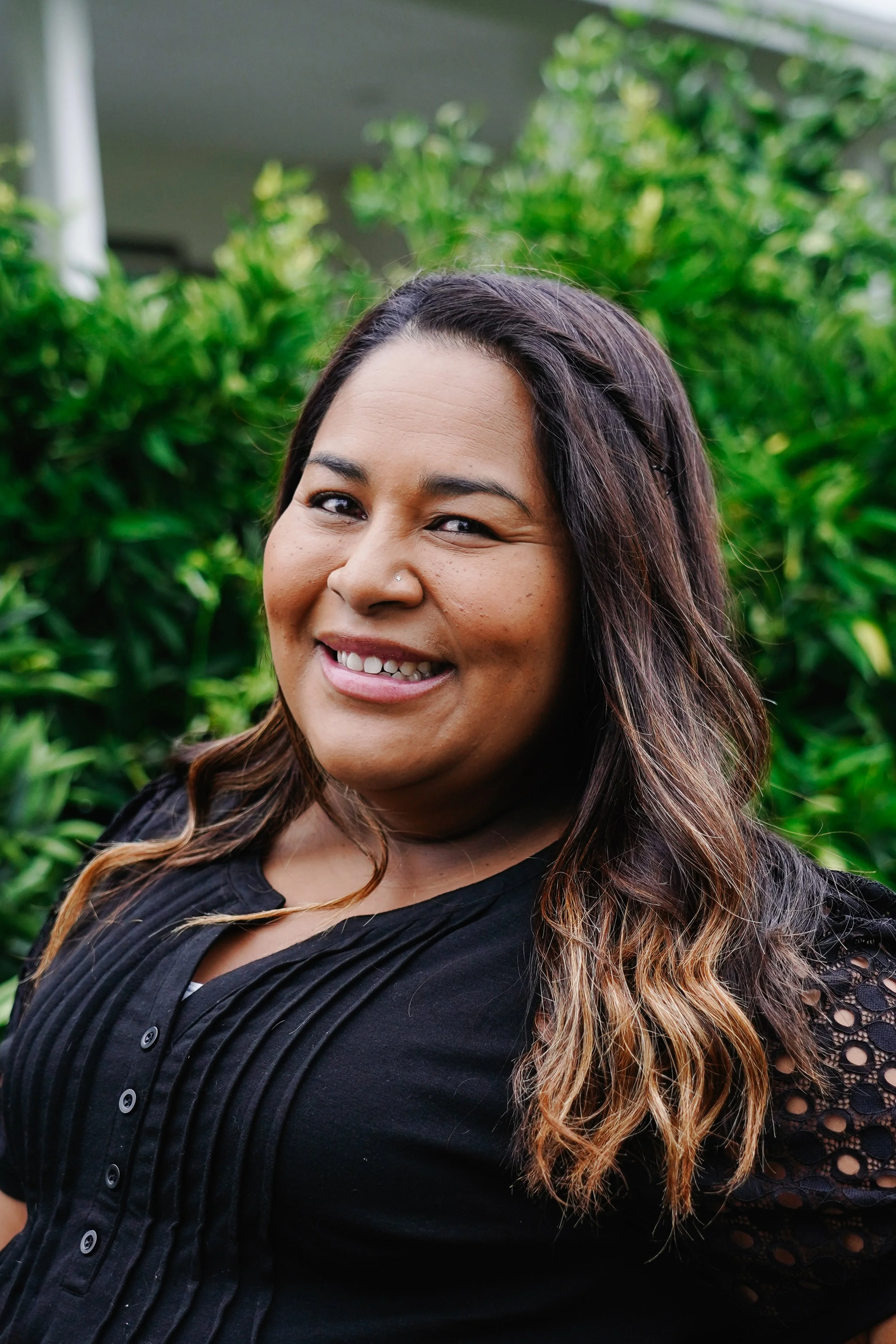 A woman with long, wavy hair smiling outdoors with green foliage in the background.