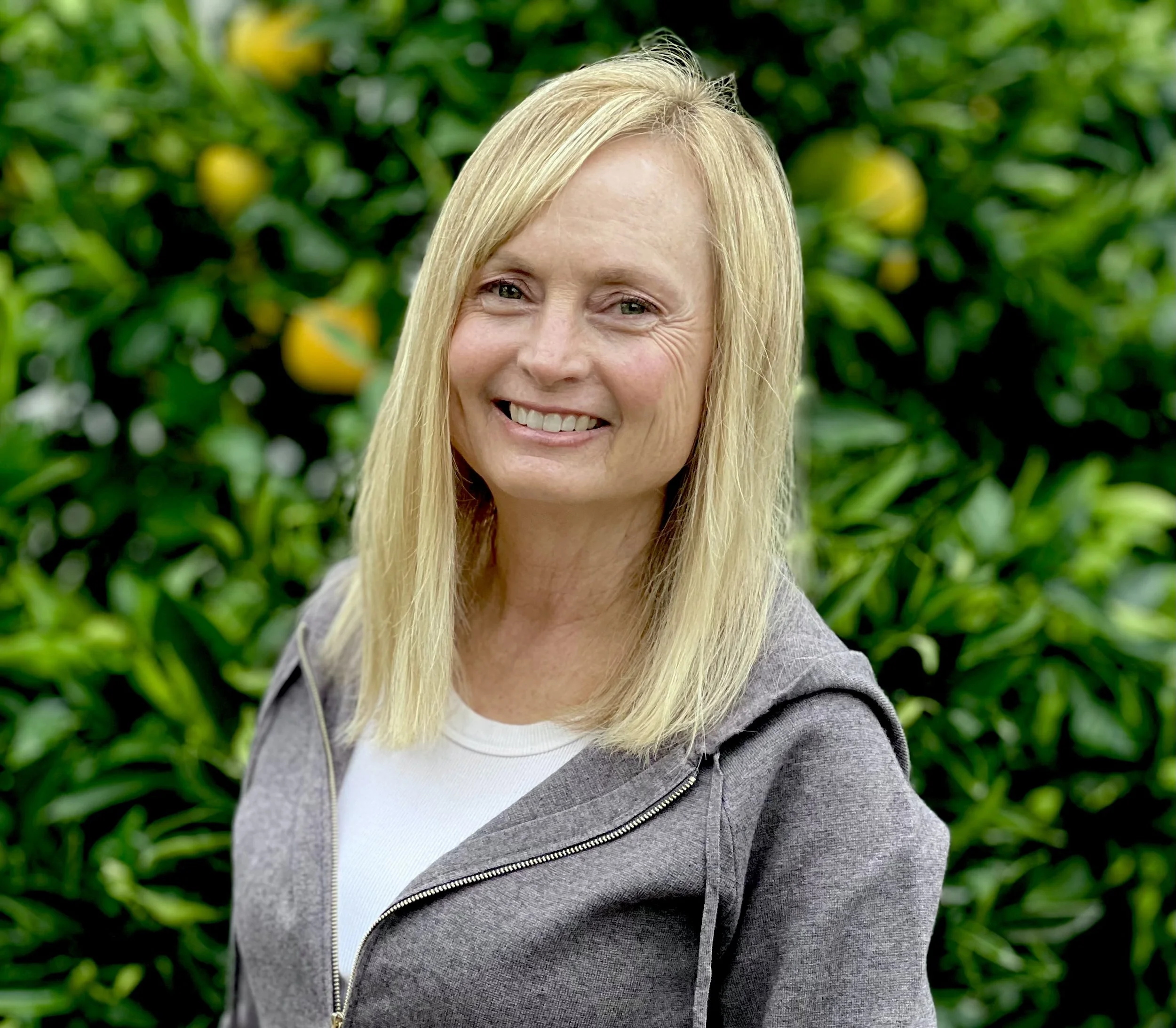 A woman with long blonde hair smiling outdoors in front of a lemon tree with yellow lemons.