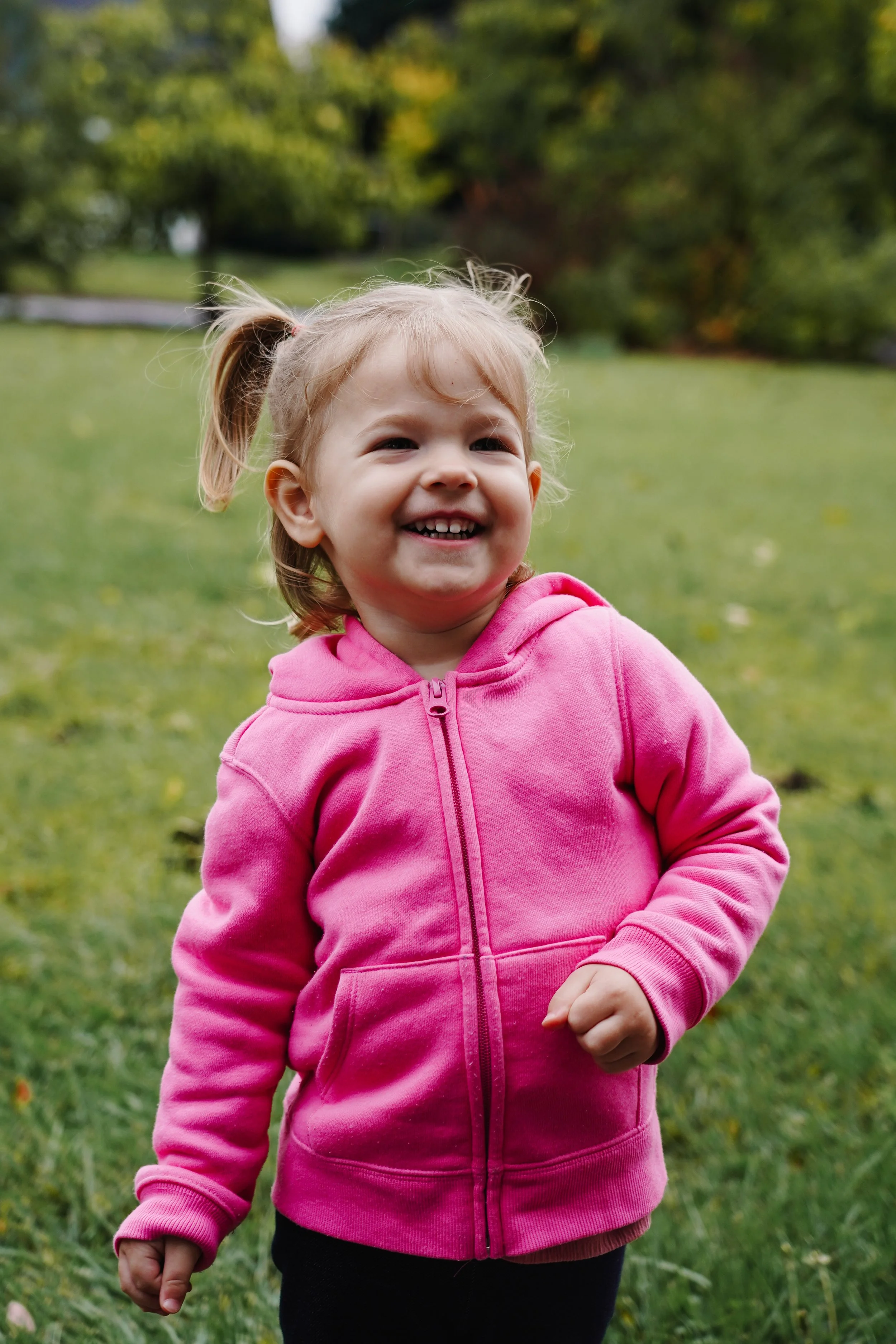 A young girl wearing a pink hoodie standing outdoors in a grassy park with trees in the background, smiling.