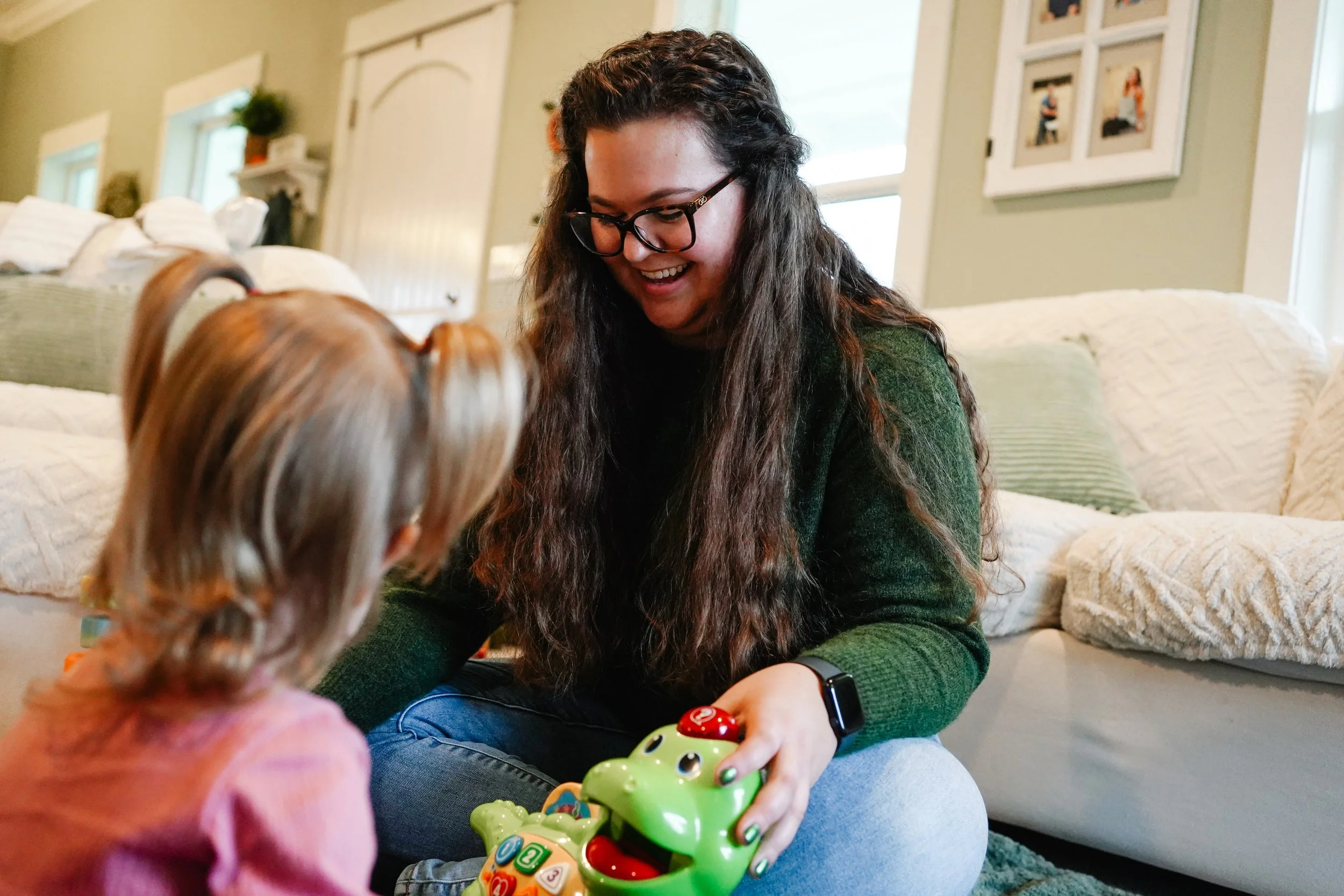 A woman with long dark hair, glasses, and wearing a green sweater smiling while playing with a young girl with blonde hair and a pink shirt on the floor of a cozy living room.
