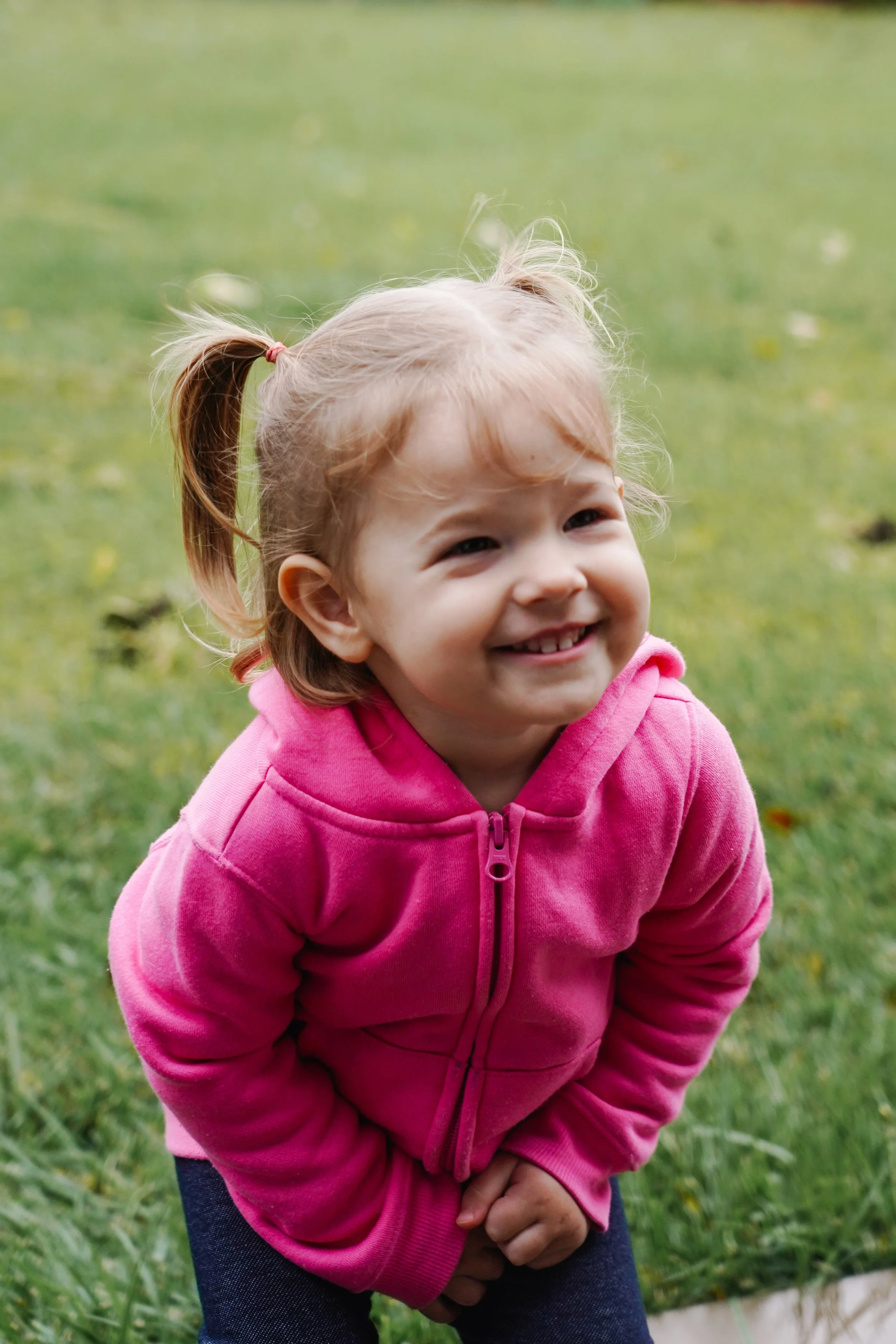 A young girl with curly hair in a ponytail, smiling and wearing a pink hoodie, standing on grass outdoors.