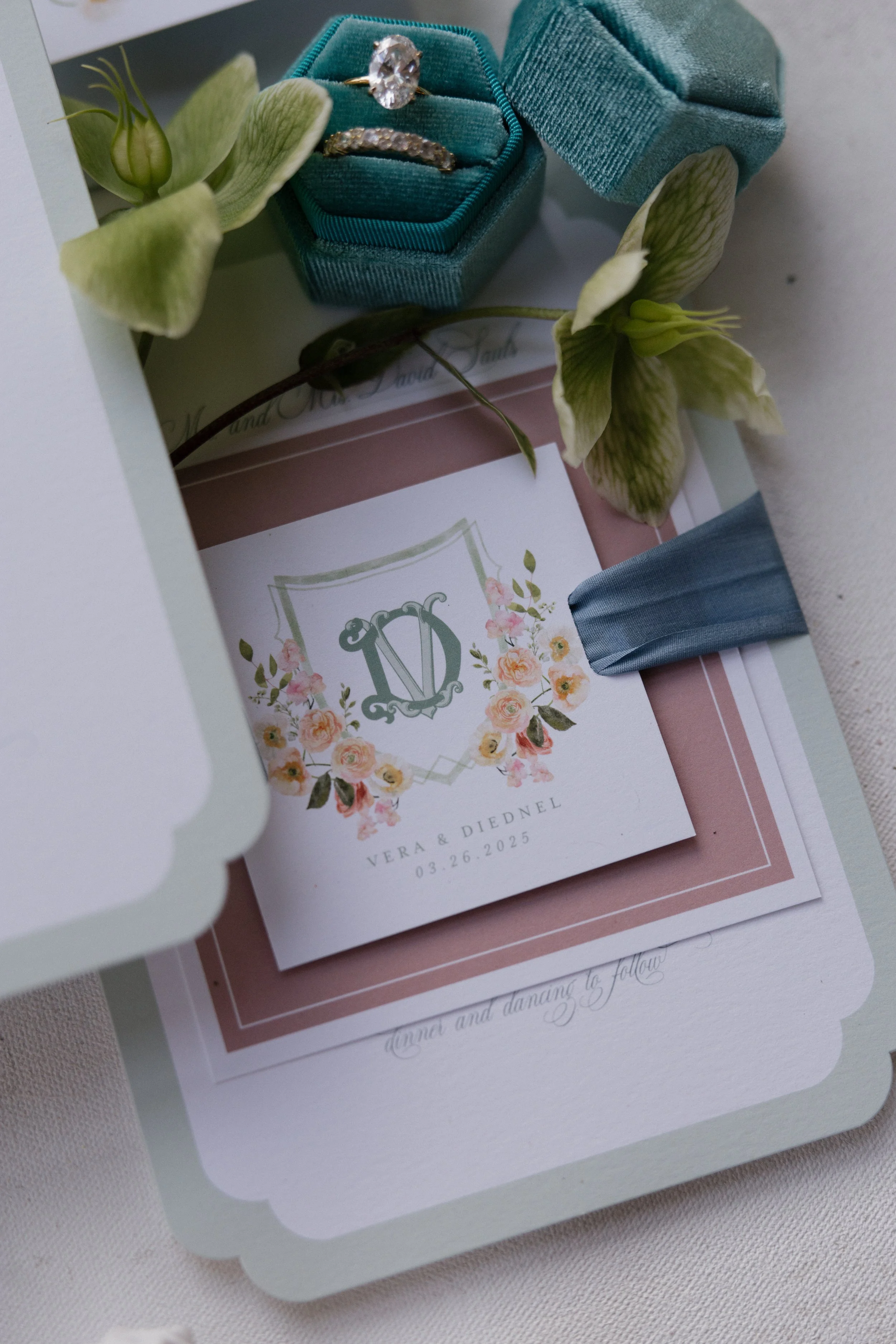 Wedding invitation with shoes, flowers, ring box, Chanel lipstick, and perfume on a floral tablecloth.