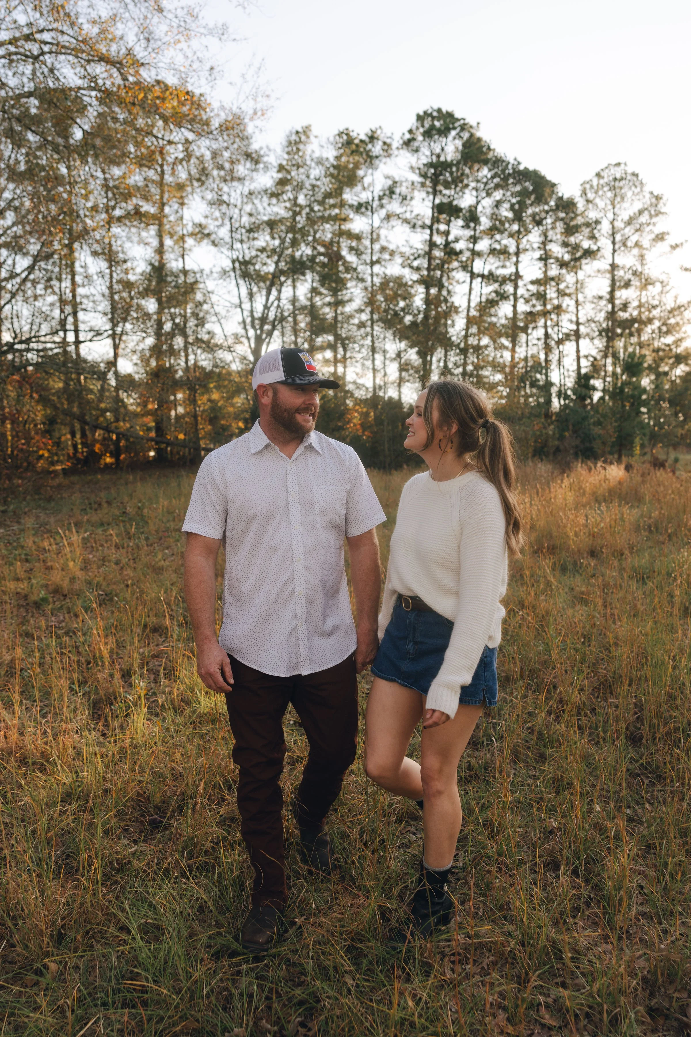 A man and woman standing in a grassy field with trees in the background, during sunset. They are holding hands and smiling at each other.