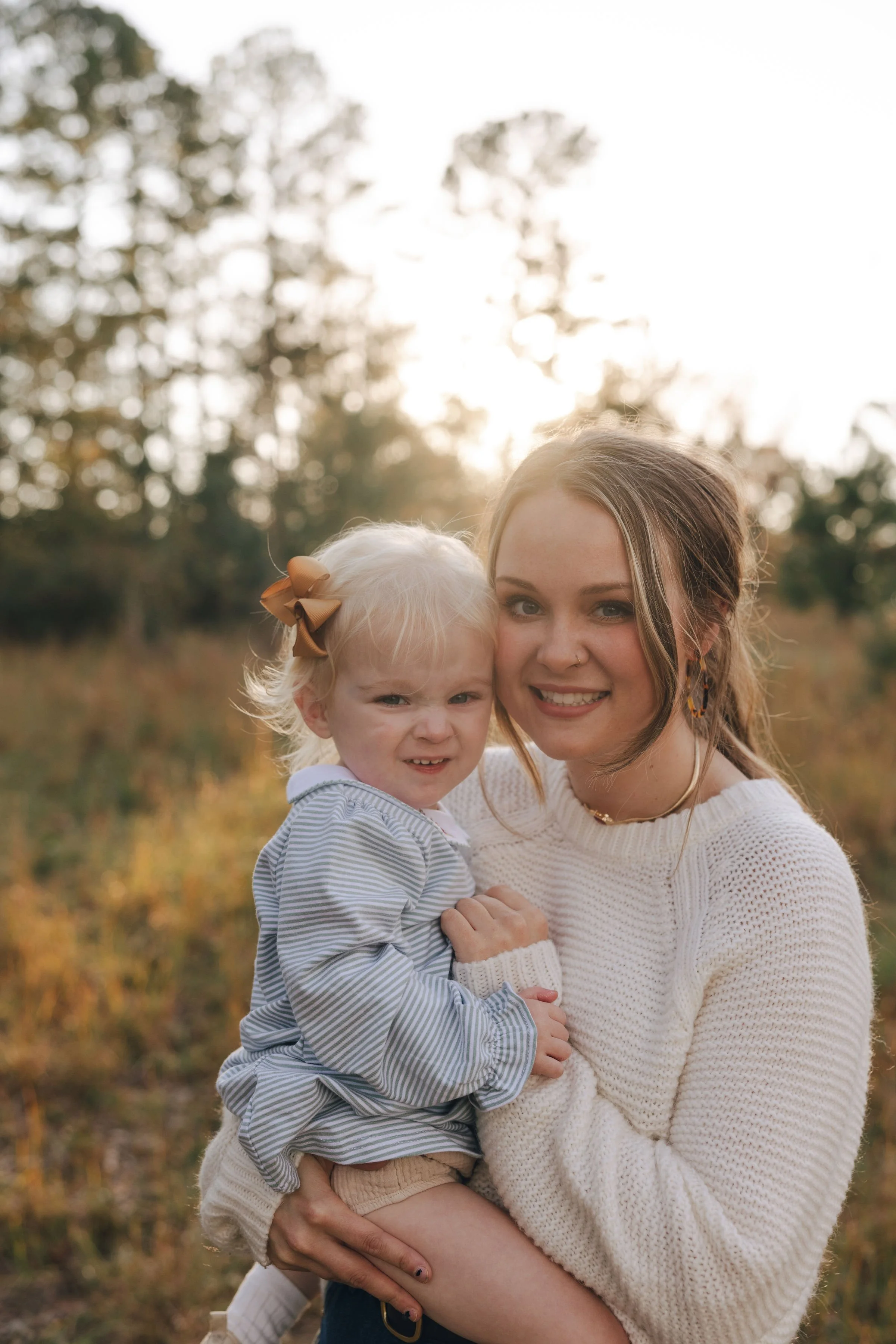 A woman holding a young child outdoors during sunset, both smiling and facing the camera, with trees and warm sunlight in the background.