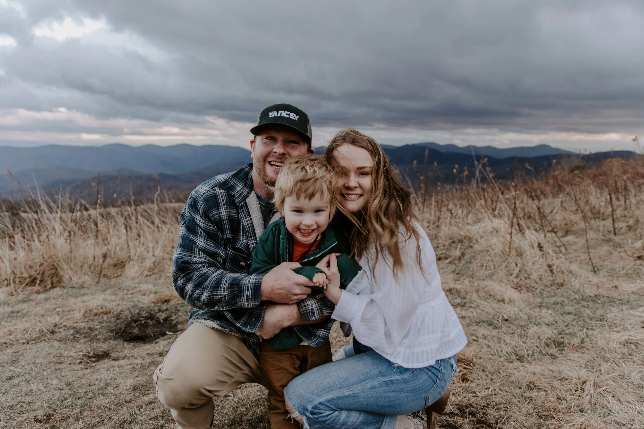 A family of three smiling and hugging outdoors with a mountain landscape and cloudy sky in the background