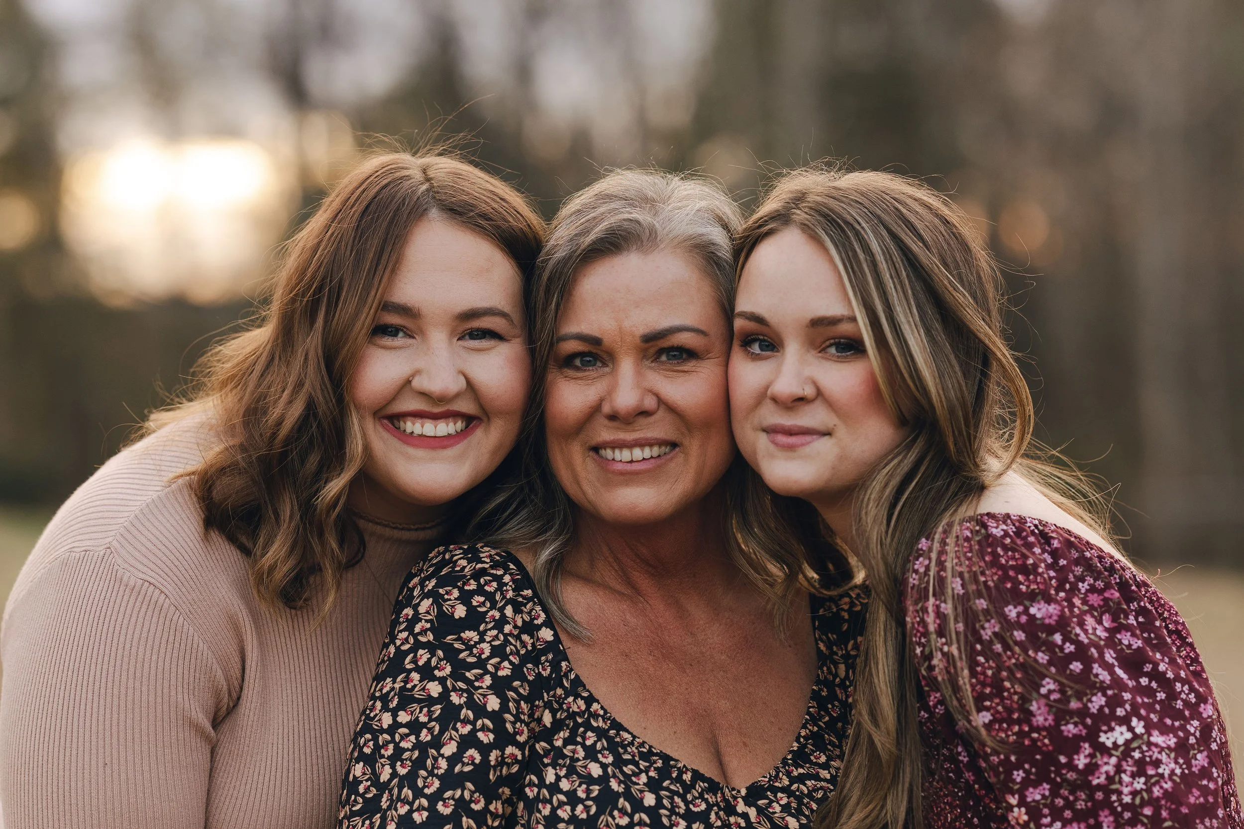 Three women smiling closely together outdoors during sunset, with blurry trees in the background.