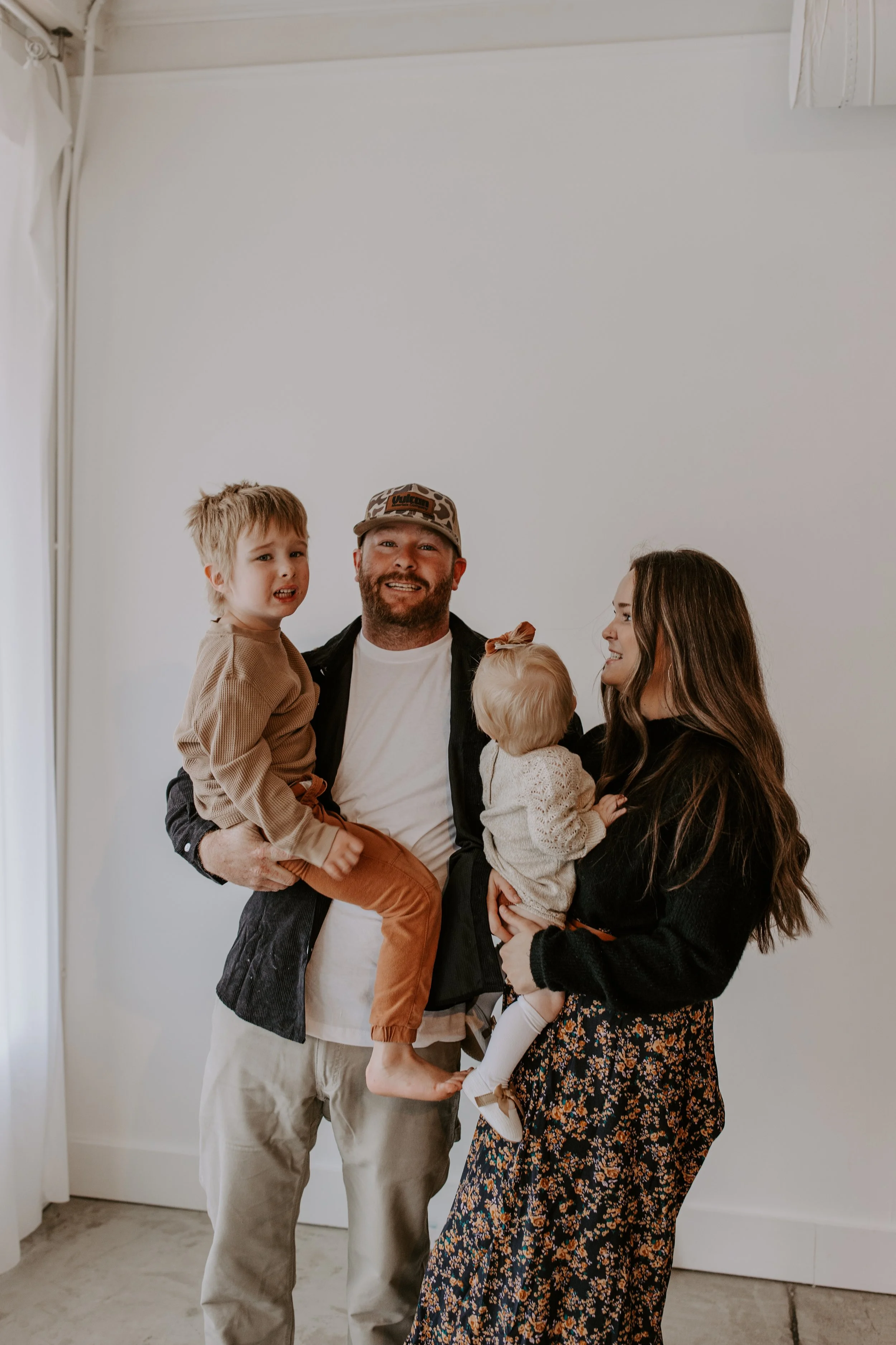 A family of four smiling and looking at the camera, standing indoors against a plain white wall. The father is holding a young boy, and the mother is holding a toddler girl.
