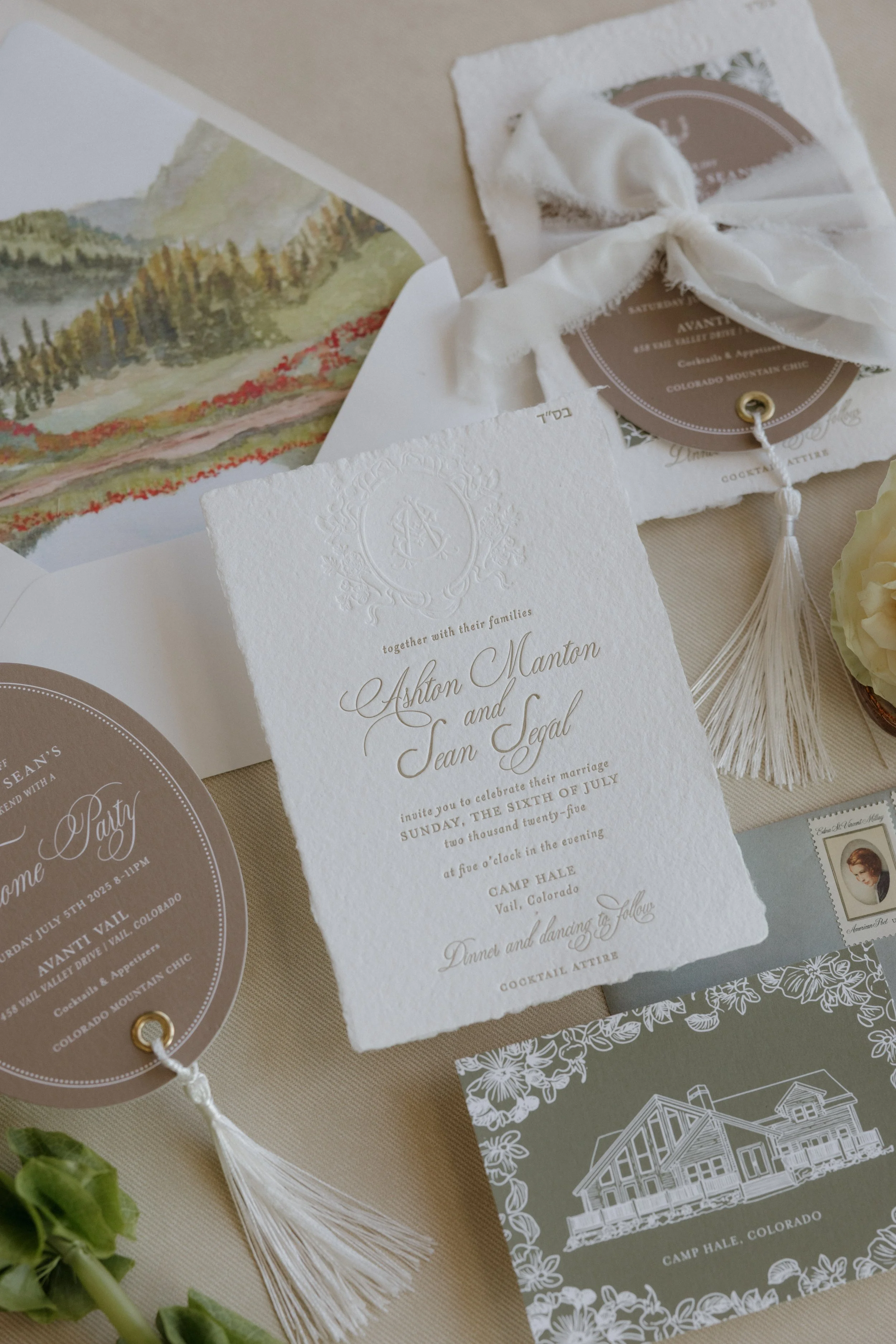 Wedding invitation suite with details of the wedding of Ashton Manton and Jean Segal, including an embossed textured card, round RSVP card, a colorful watercolor map of Colorado, and decorative elements on a beige surface.
