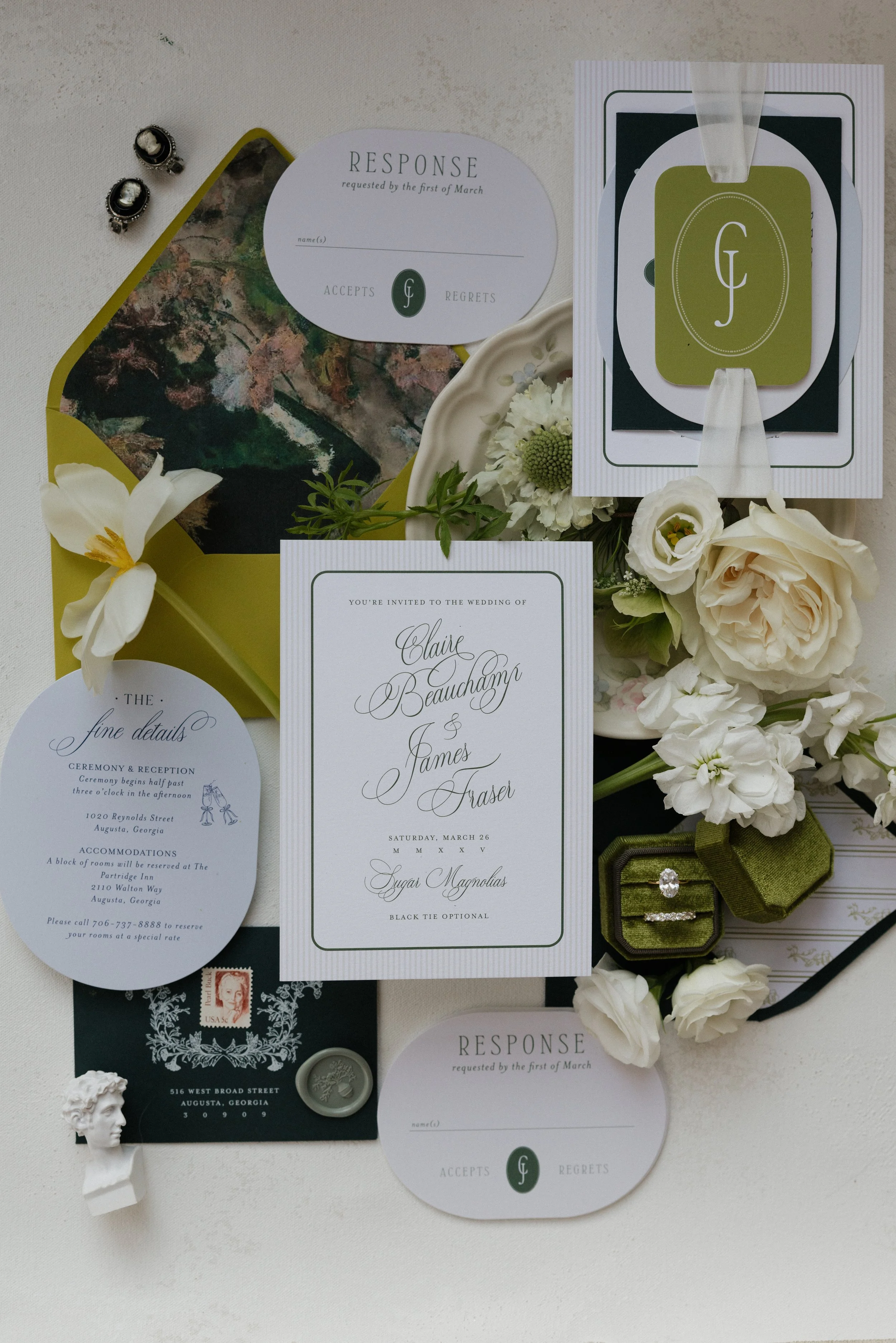 Wedding invitation and RSVP cards with white and green accents, surrounded by flowers, rings, and decorative items on a white surface.