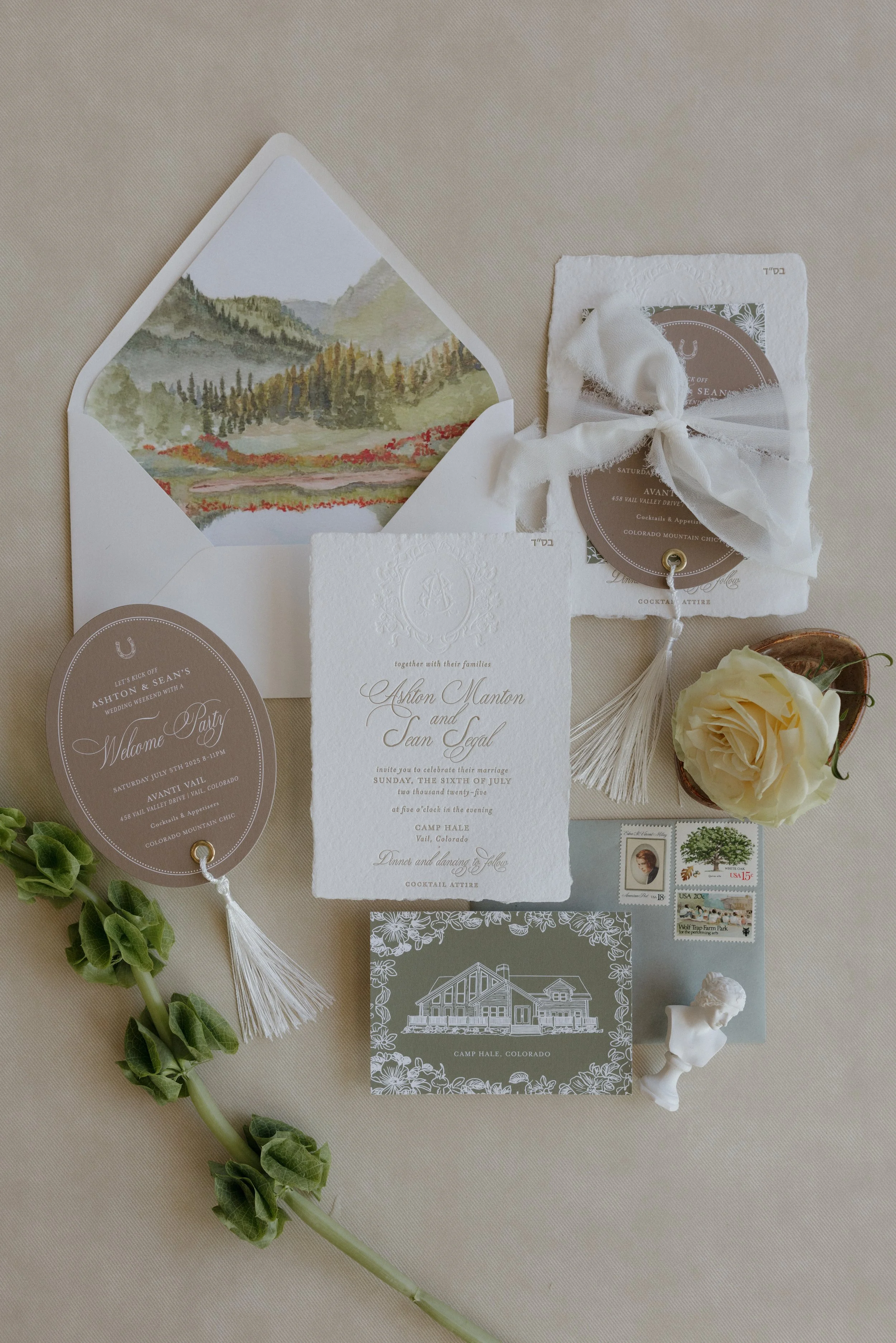 Wedding invitation suite with a white textured card, a round welcome party card, an address card with a bow, floral stamps, a white rose, a watercolor mountain landscape inside an envelope, and green foliage, all arranged on a light-colored surface.