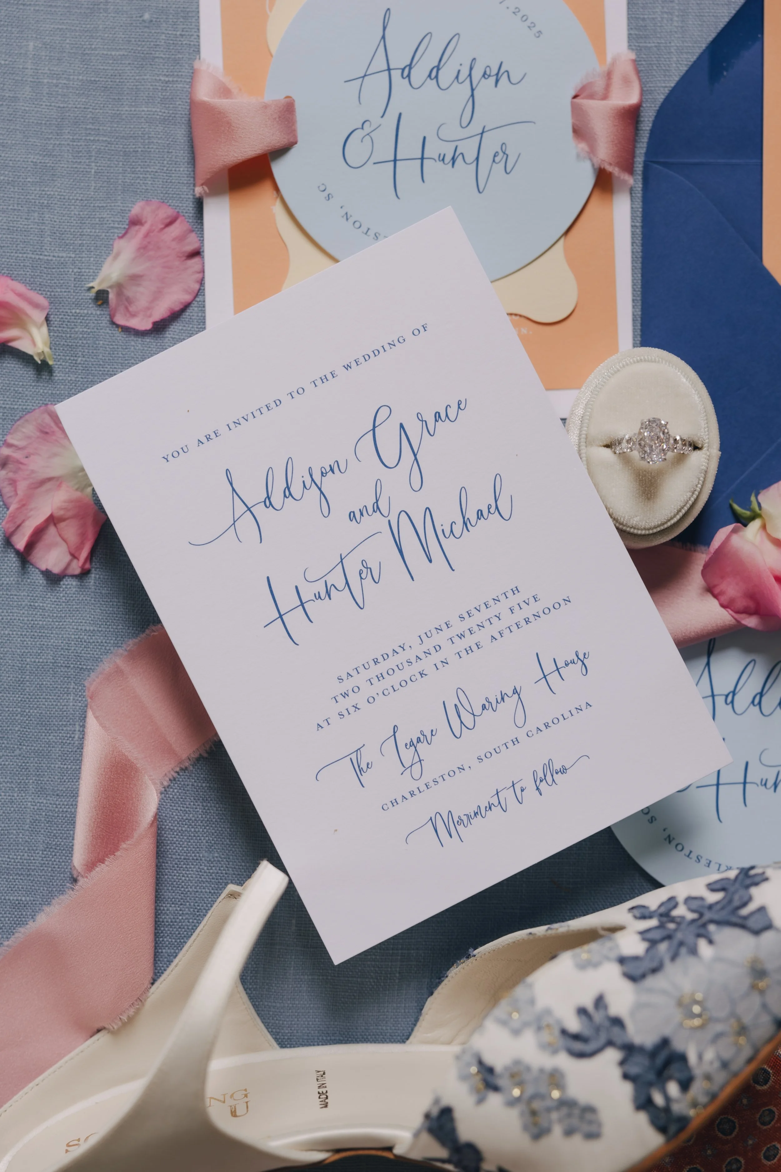 Wedding invitation for Addison Grace and Hunter Michael with an engagement ring and pink flower petals on a blue and peach background.