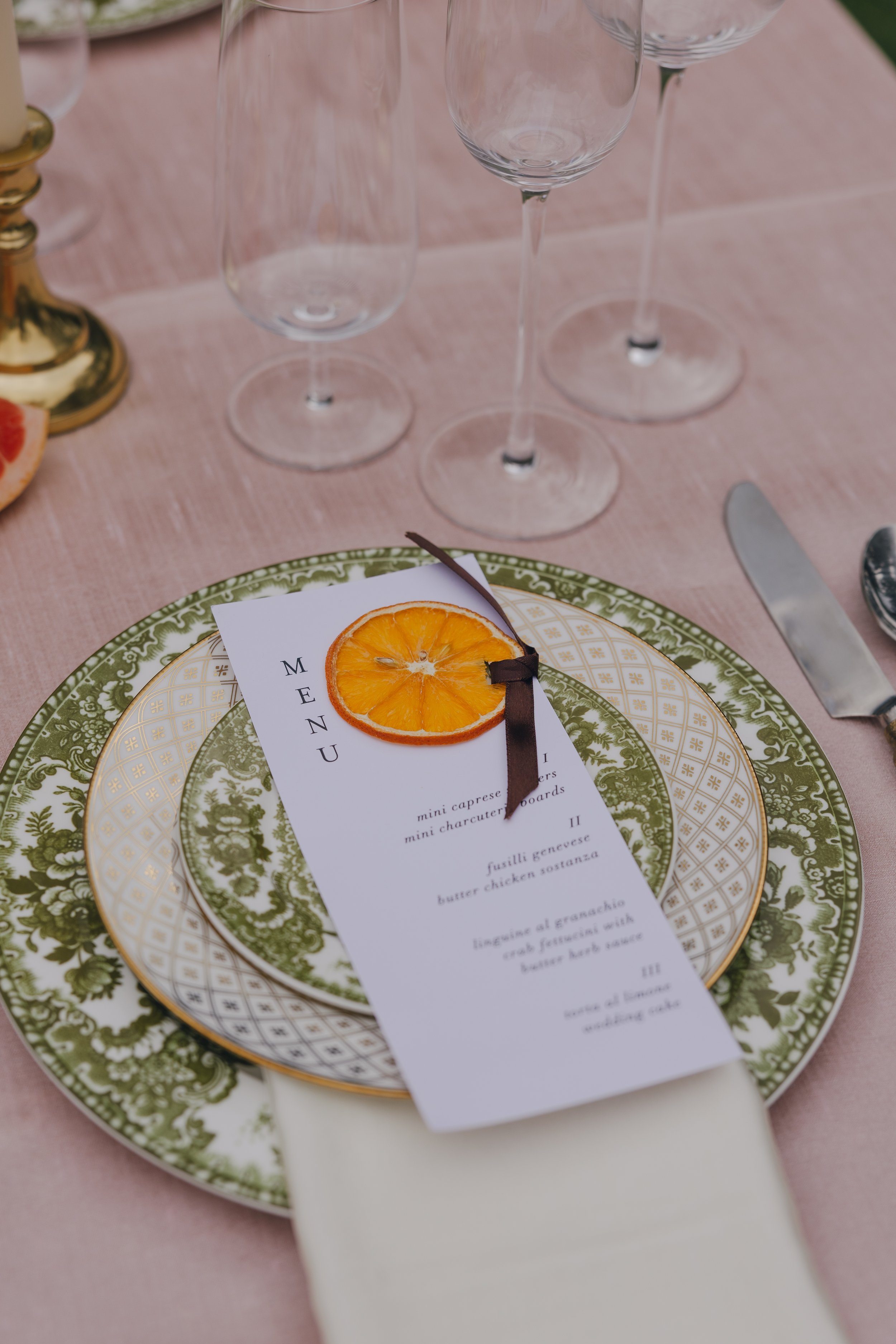 Elegant table setting with a menu card topped with dried orange, placed on decorative plates, with empty wine glasses and a pink tablecloth.