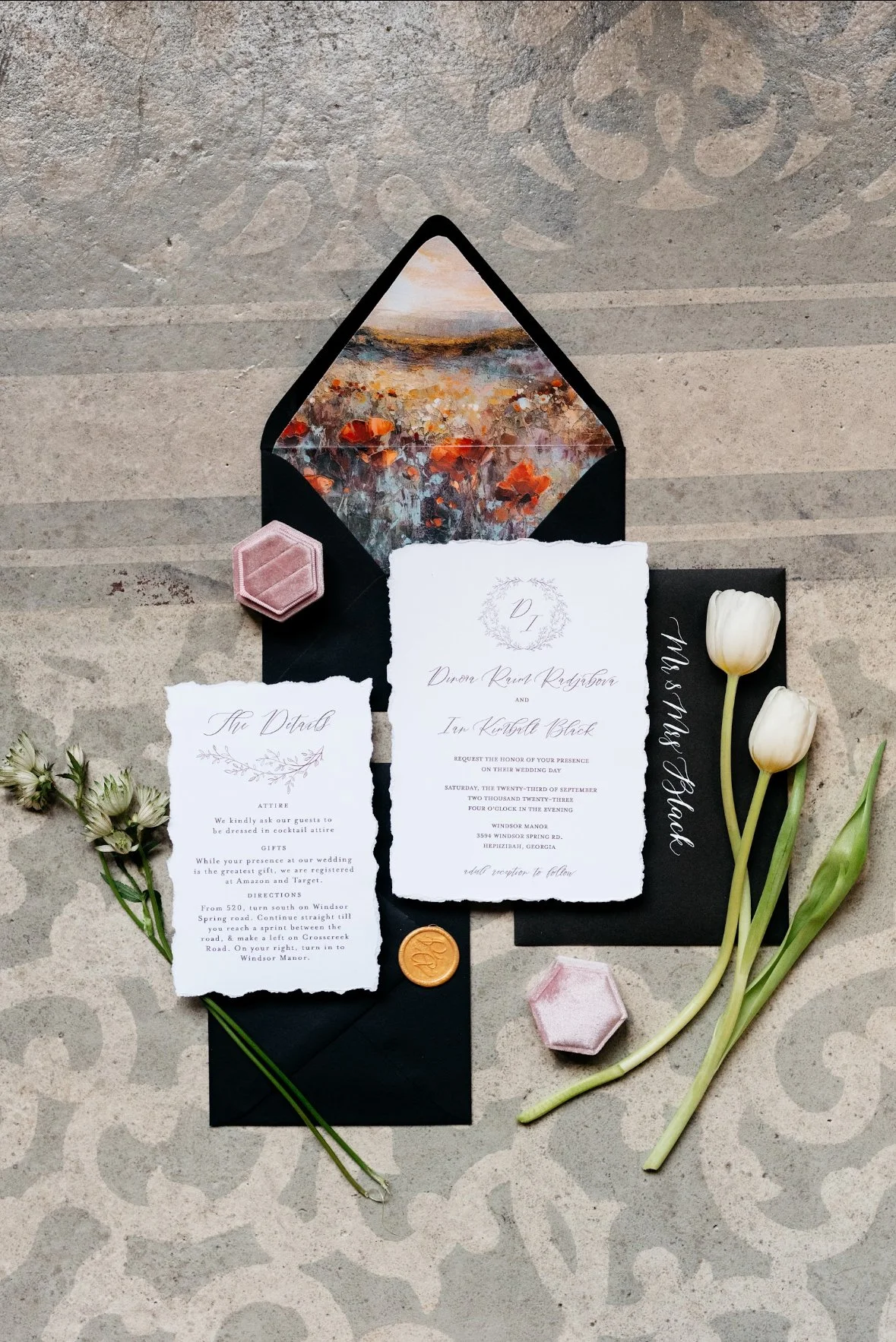 Wedding invitation suite with a black envelope, a handwritten RSVP card, and a detail card, accompanied by white tulips, an orange wax seal, and pink velvet jewelry boxes, all set on a decorative surface.