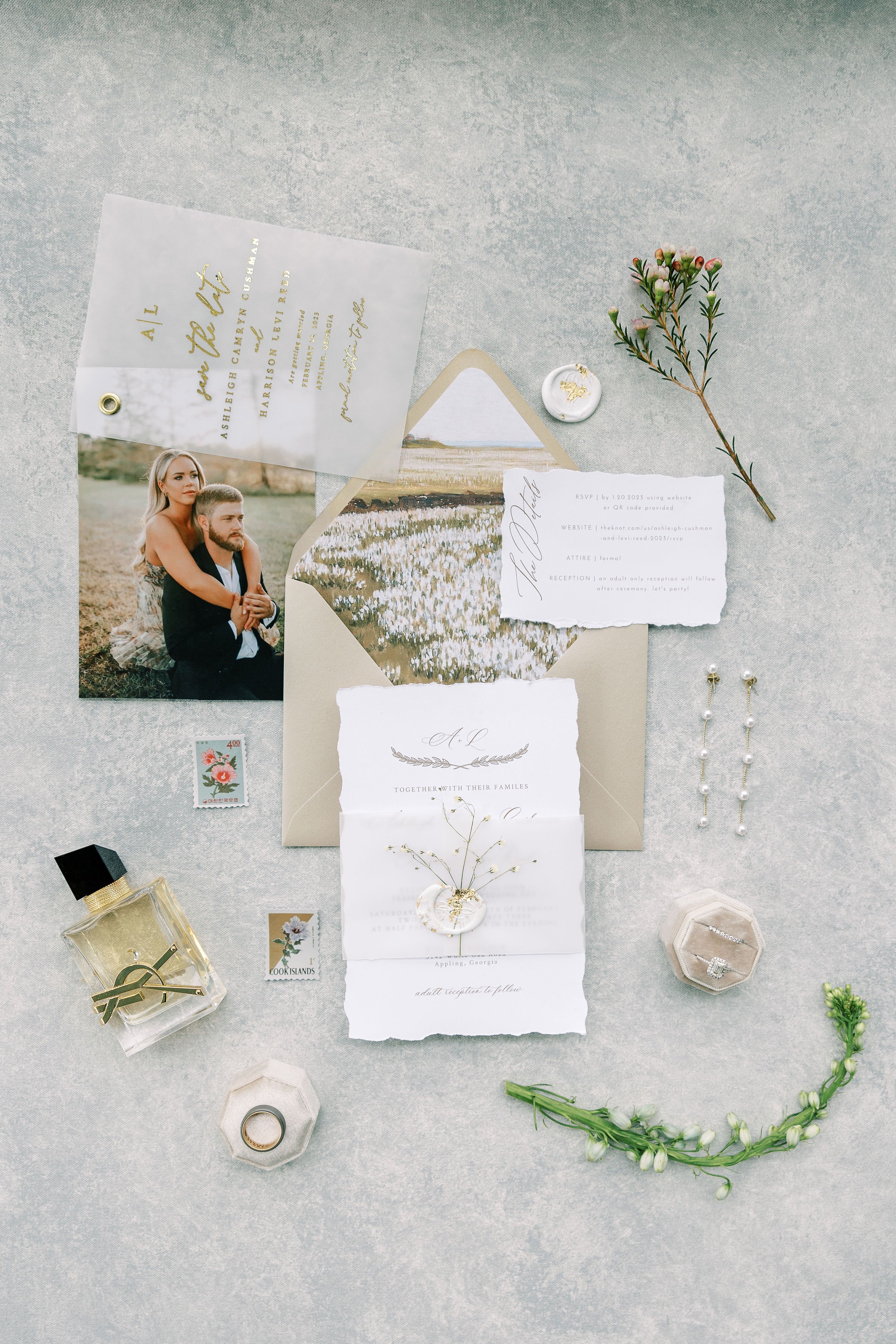 Wedding invitation suite with photography, floral accents, jewelry, and small decorative items on a grey surface.