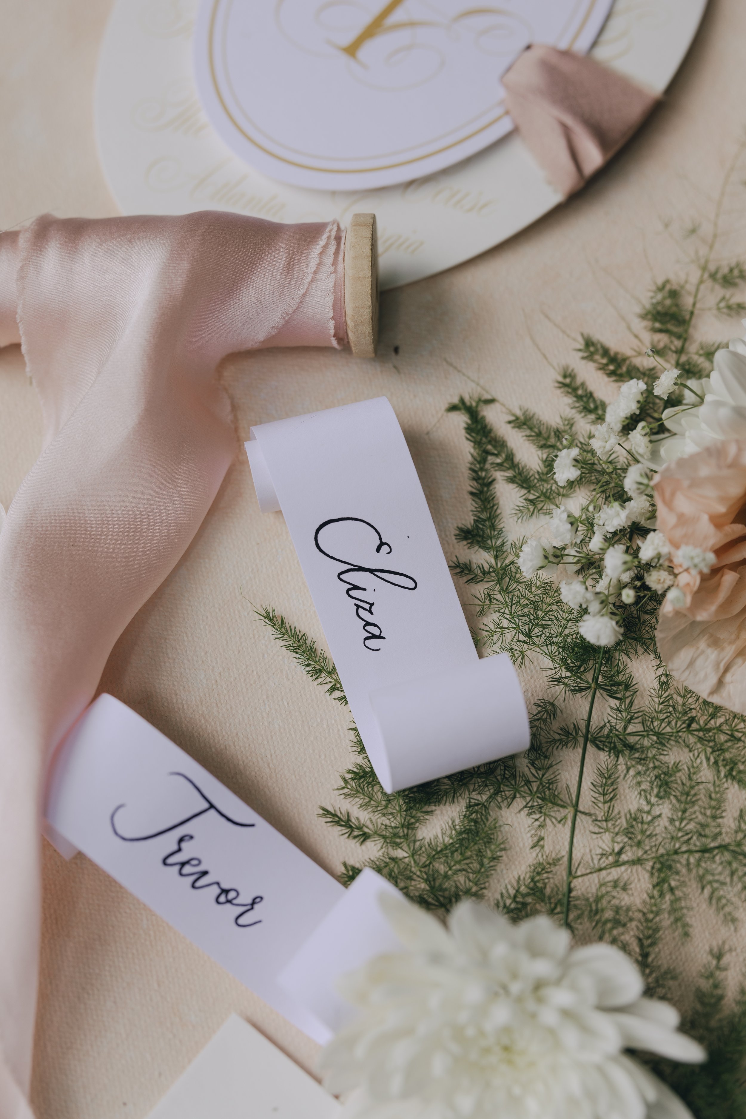 Wedding table decor featuring pink satin ribbons, flower name tags reading 'Chiara' and 'Javier', white flowers, and green foliage.