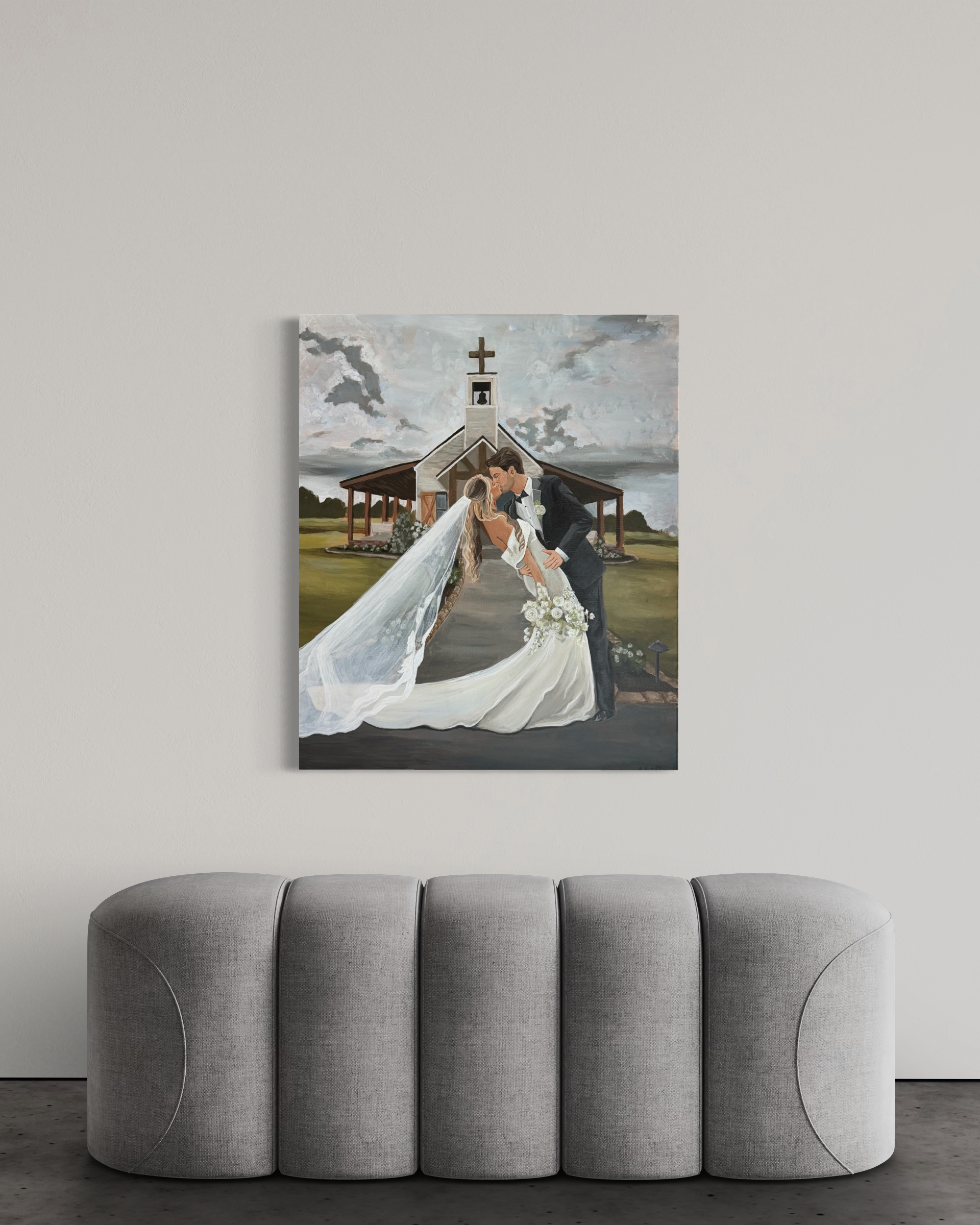 Painting of a bride and groom kissing in front of a church, with a cloudy sky in the background, hung on a white wall above a grey sofa.
