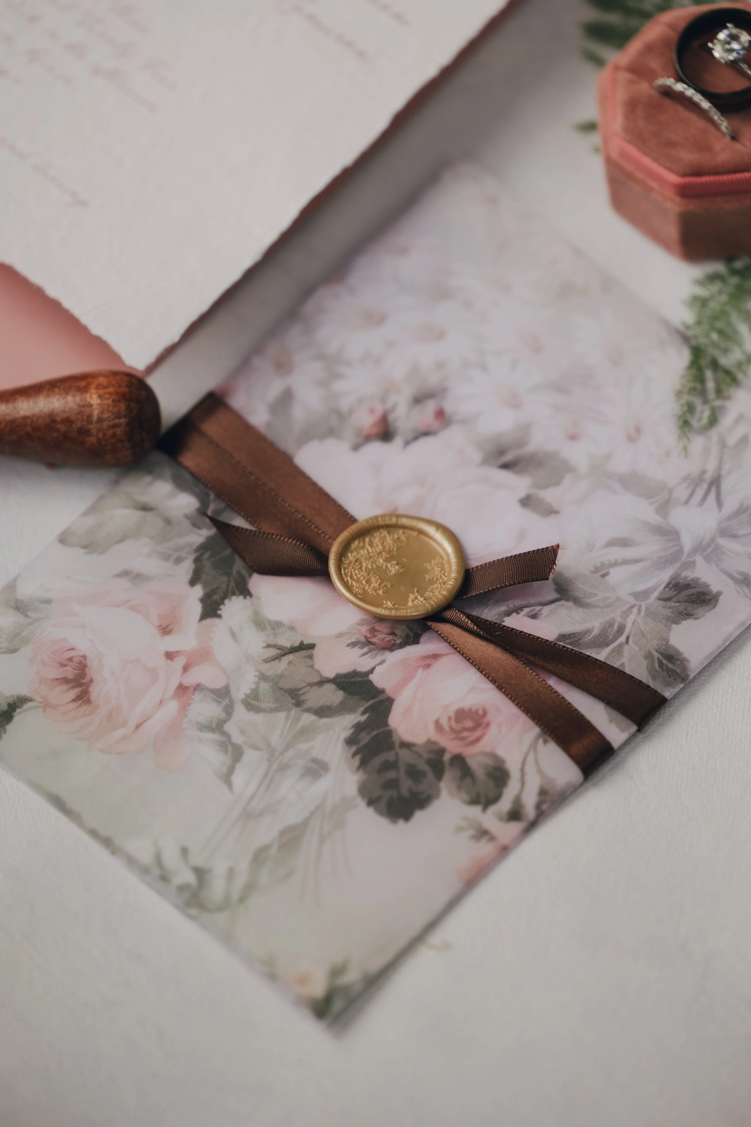 A wedding invitation with floral design, tied with a brown ribbon and wax seal, accompanied by rings and a letter on a white surface.