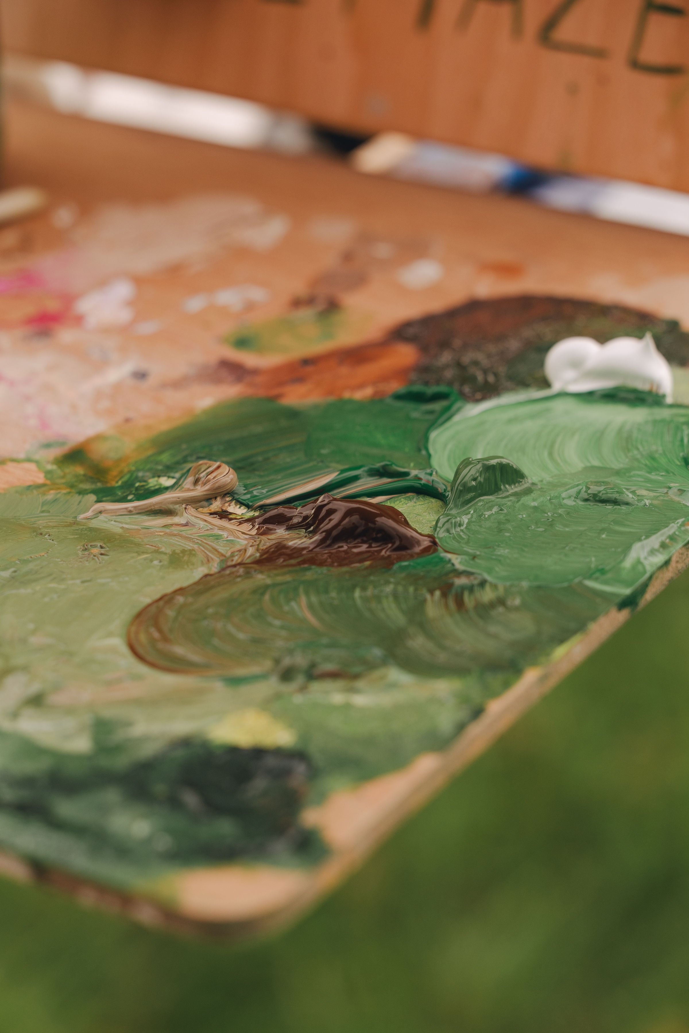 Close-up of an artist's palette with thick, textured acrylic paint in shades of green, brown, white, and hints of other colors.