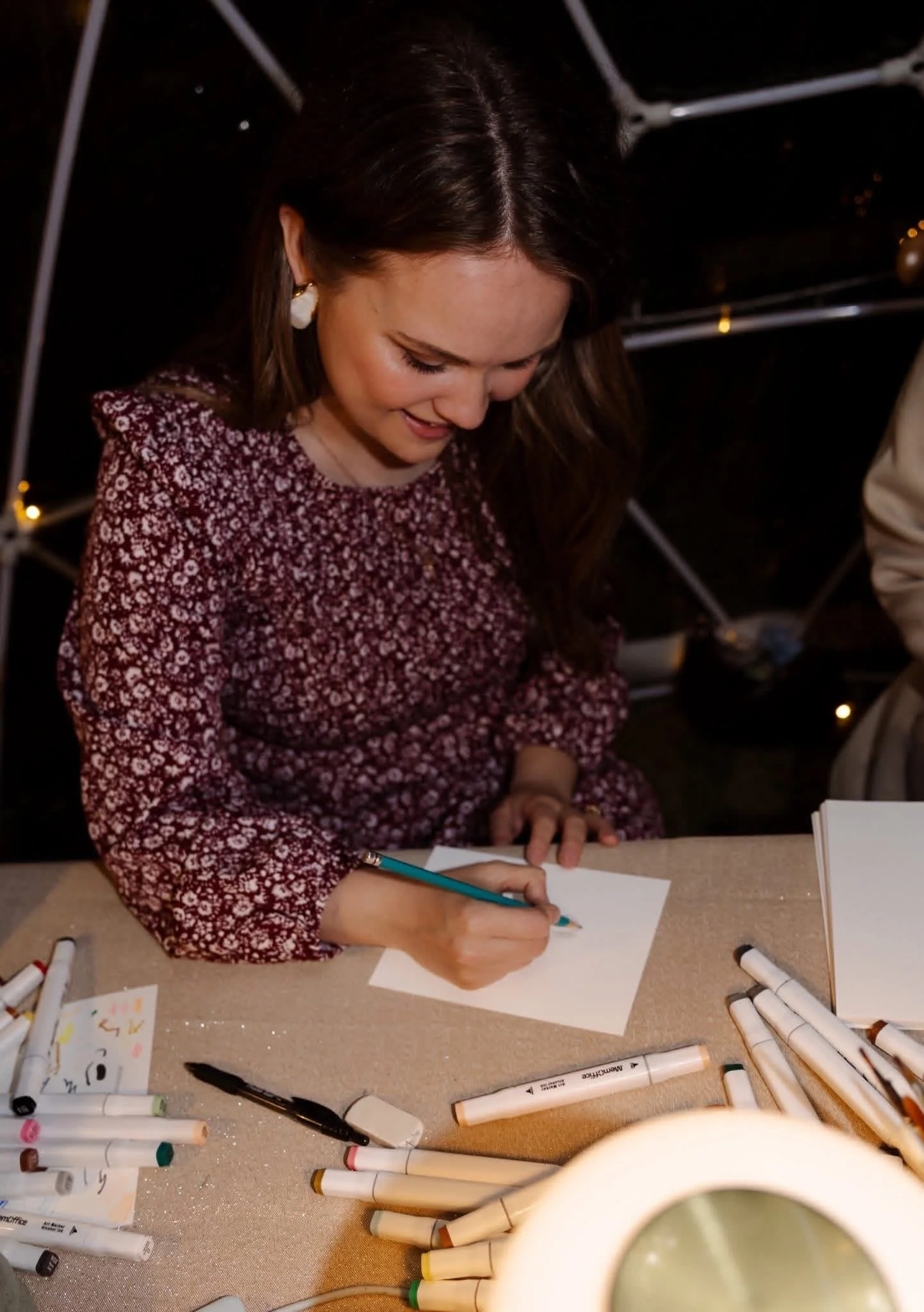 A woman with long brown hair, wearing a red floral dress, smiling and drawing with a marker on a piece of paper. She's sitting at a table with various markers scattered around, under warm lighting.