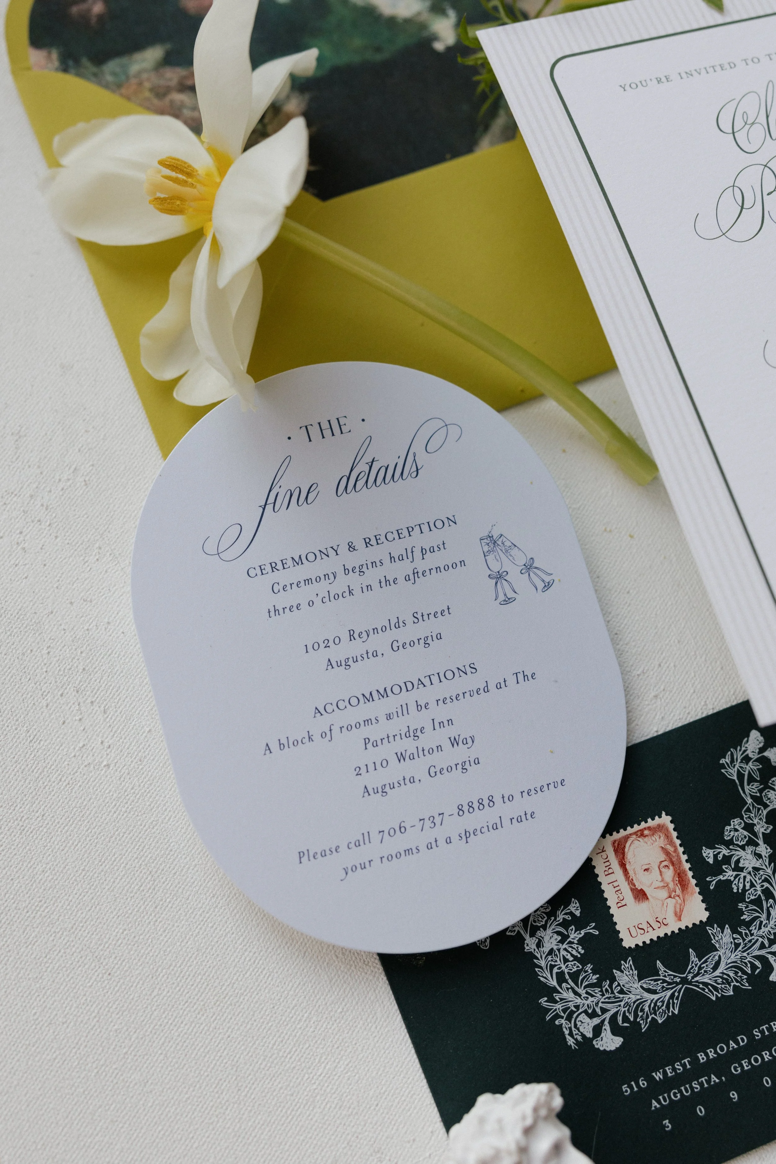 Wedding invitation with details about the ceremony and reception, addressing in Augusta, Georgia, along with an RSVP phone number, placed next to a white flower and a black envelope with a stamp.
