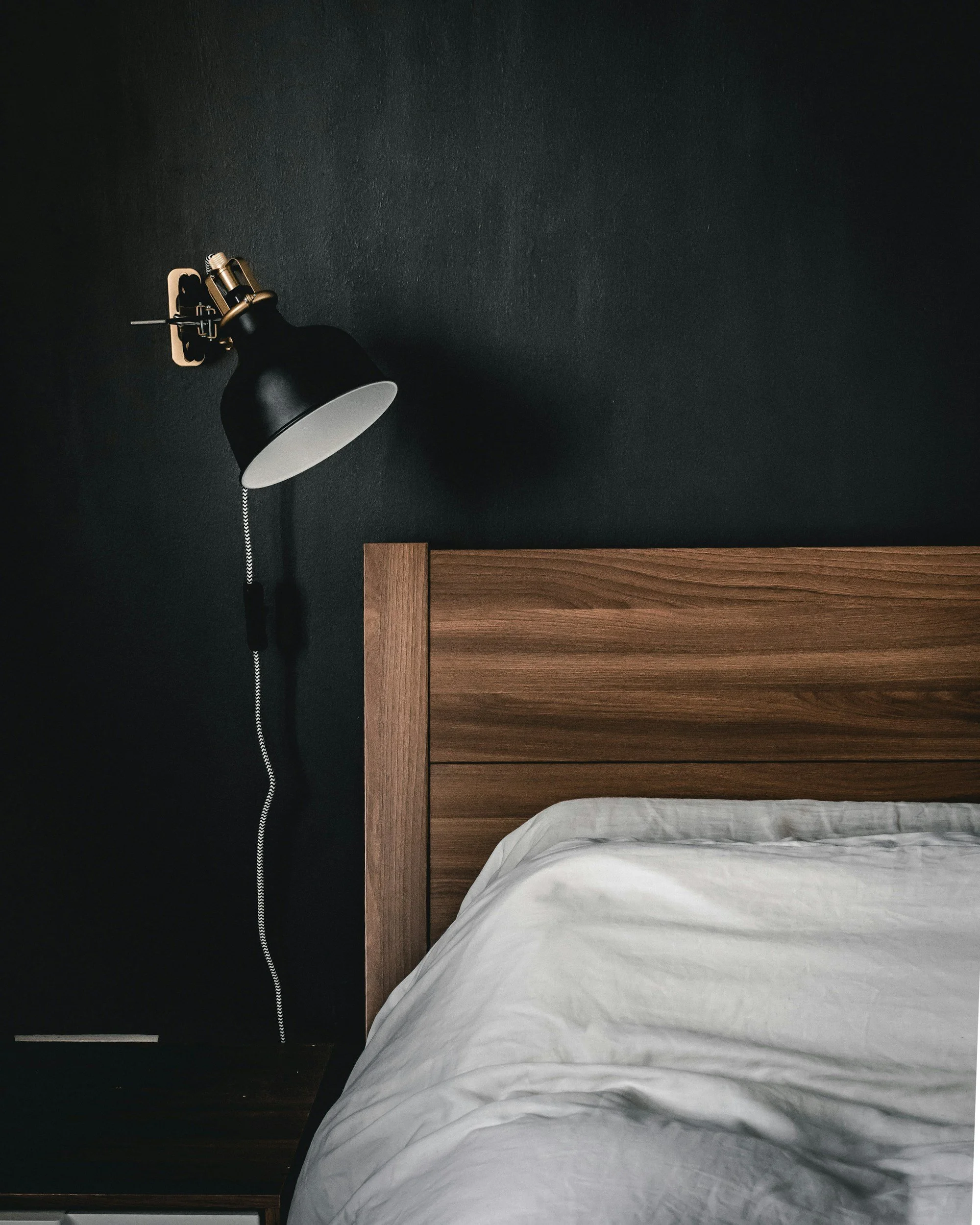 Close-up of a bedroom with a black wall, a black wall-mounted lamp, a wooden headboard, and a white bed sheet.