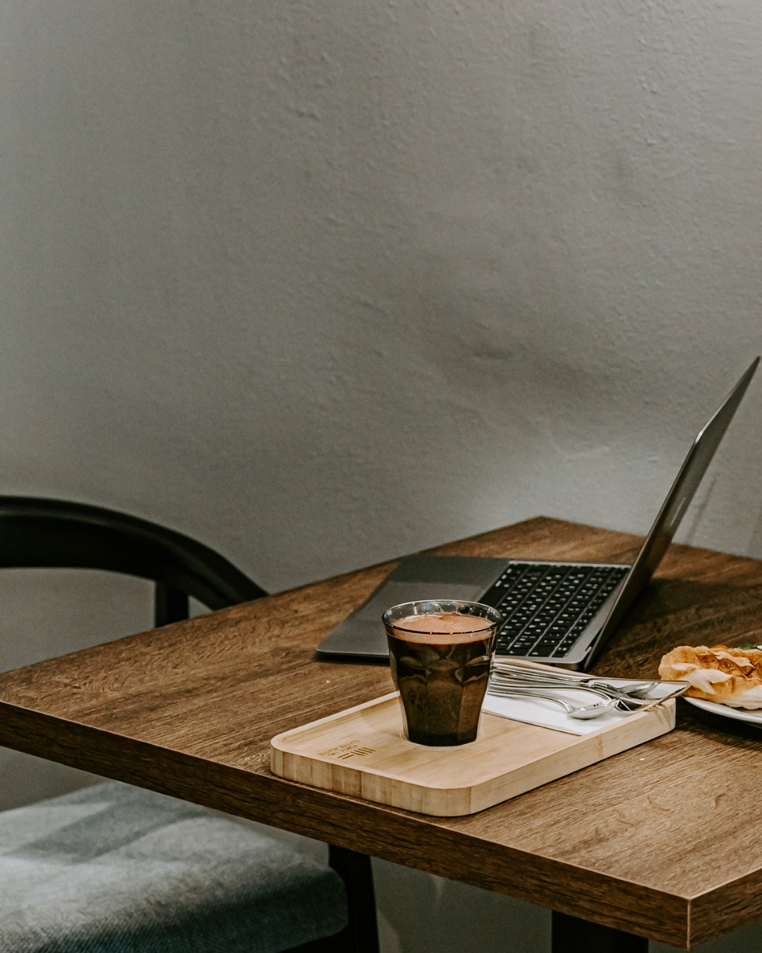 A wooden table with a laptop, a glass of dark soda on a wooden tray, and a plate with a pastry.