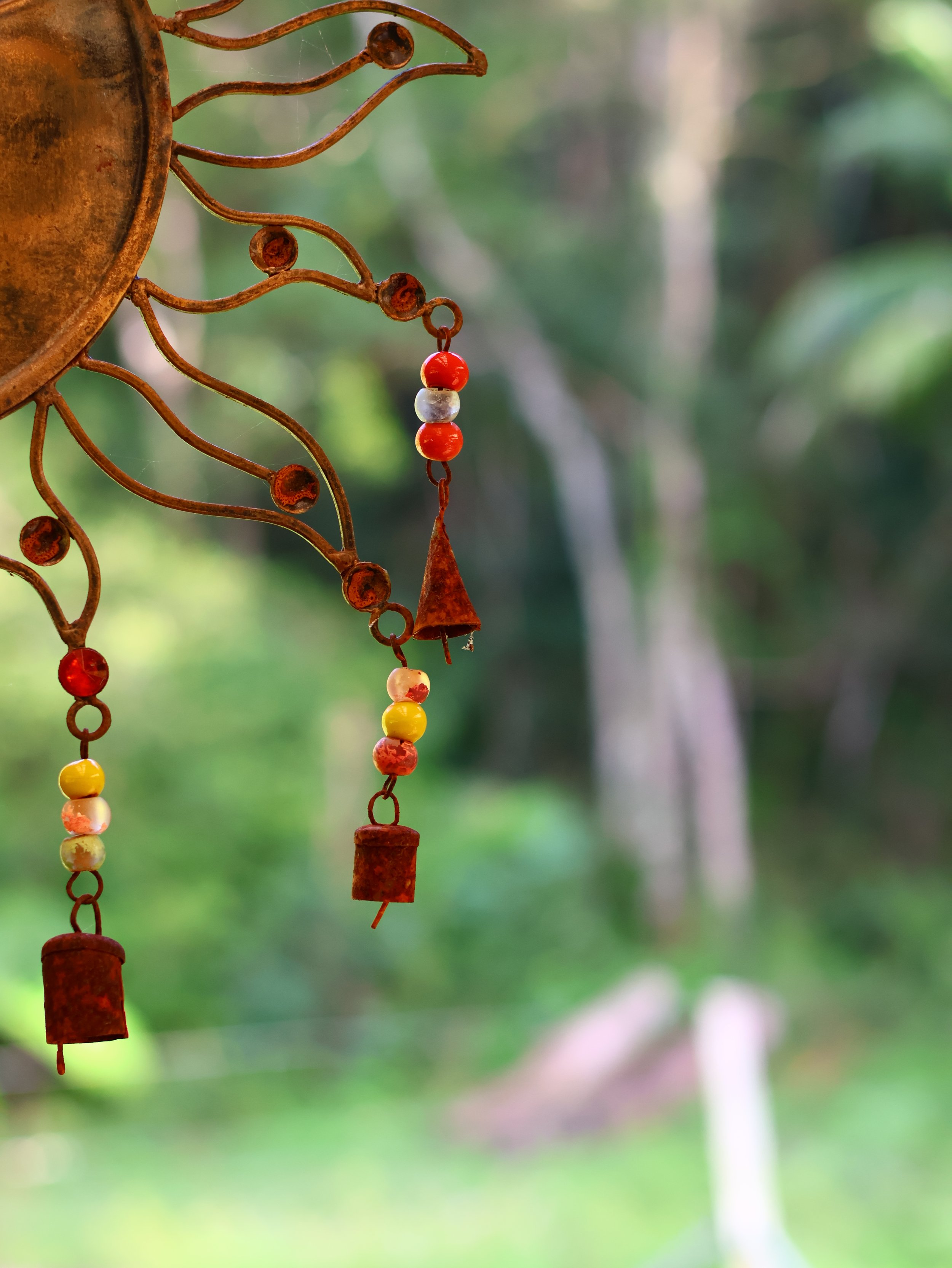 Close-up of a rusty metal wind chime with colorful beads hanging against a blurred green outdoor background.
