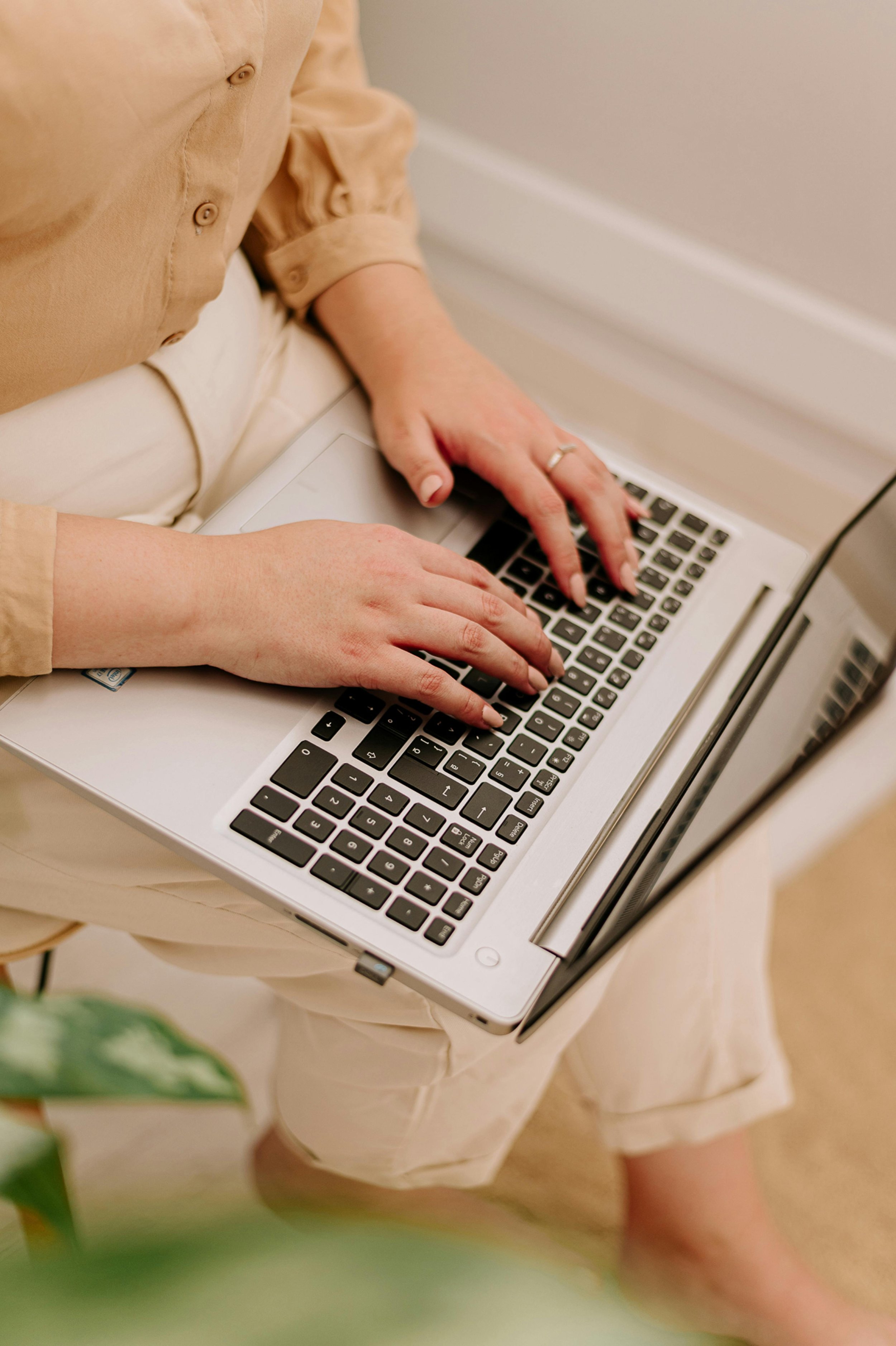 Person sitting with a laptop on their lap, typing on the keyboard. Hand with a ring on the ring finger is visible, dressed in a beige shirt.