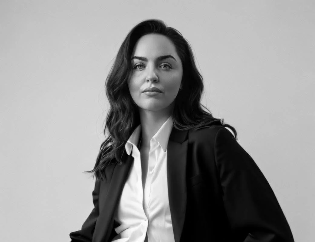 A woman with dark, wavy hair wears a black blazer over a white collared shirt, gazing confidently at the camera against a plain background.